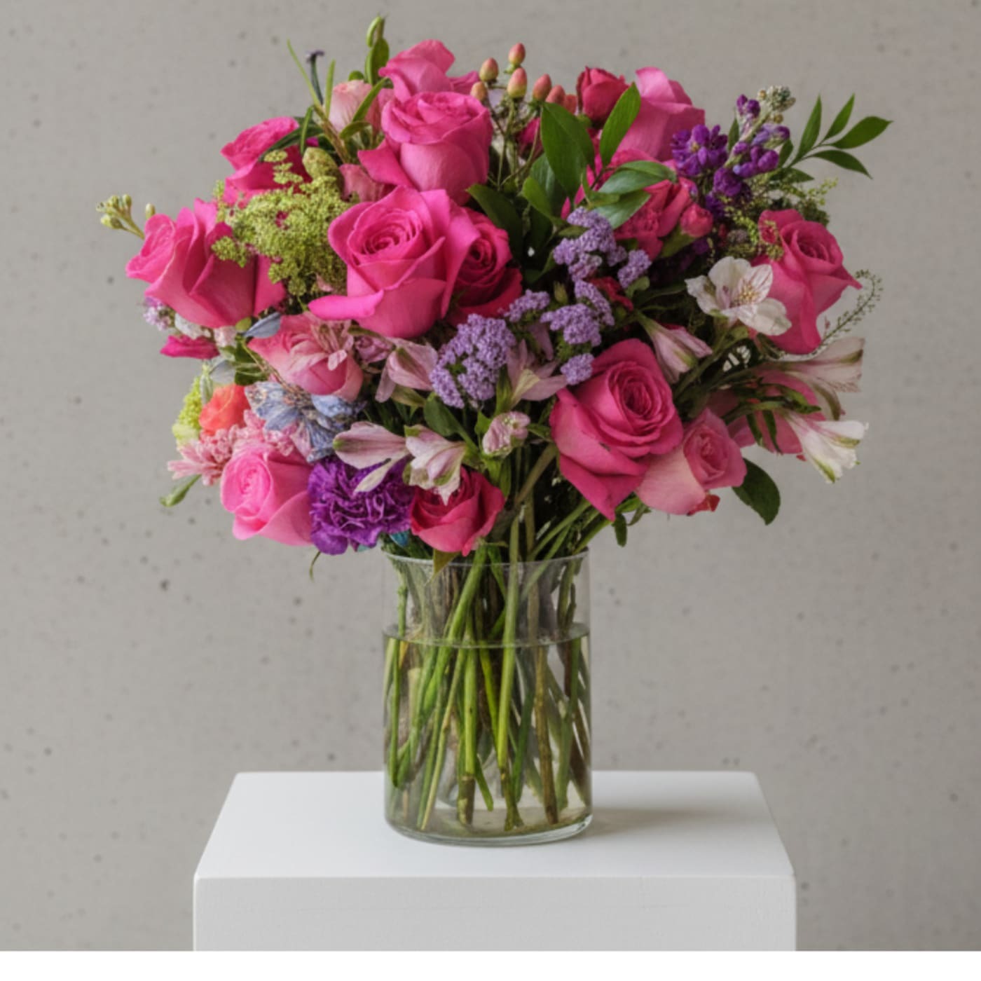 Bursting with energy and charm, this arrangement showcases bold fuchsia roses paired with soft lavender accents, delicate alstroemeria, and textured seasonal blooms. Designed in a clear glass cylinder vase, it’s the perfect choice for birthdays, anniversaries, or to simply brighten someone’s day with a joyful pop of color.