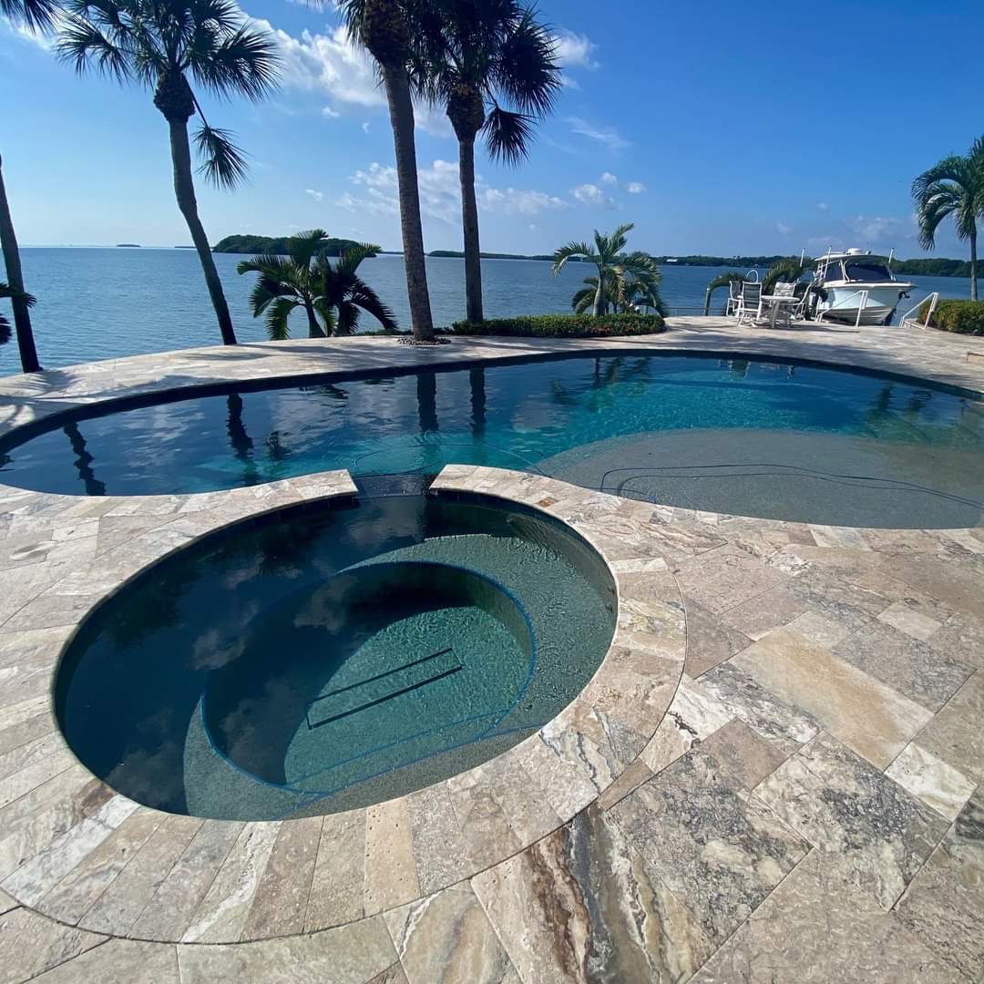 Waterfront residential swimming pool with an attached round spa, stone paver decking, palm trees, and scenic bay views.