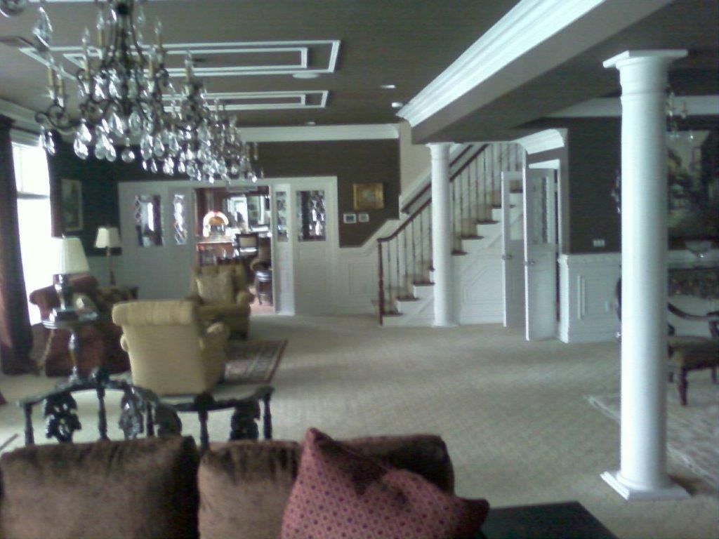 A spacious, elegant living area with chandeliers, white columns, and a staircase. Plush seating, decorative lighting, and open layout create a luxurious atmosphere ideal for relaxing and entertaining guests.