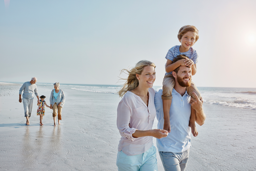 A family enjoys a sunny day on the beach. A man carries a smiling child on his shoulders, walking alongside a woman. An elderly couple and child stroll behind them.