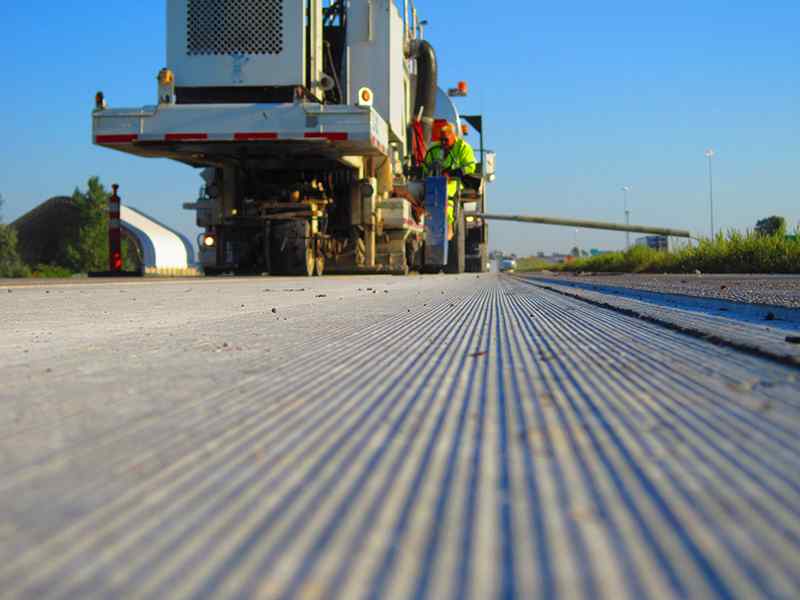 Low-angle close-up emphasizing the precise, uniform longitudinal grooves created by a diamond grinding machine on concrete pavement. In the background, the rear of a DSI grinding rig with an operator in high-visibility gear moves along the roadway under clear blue skies.