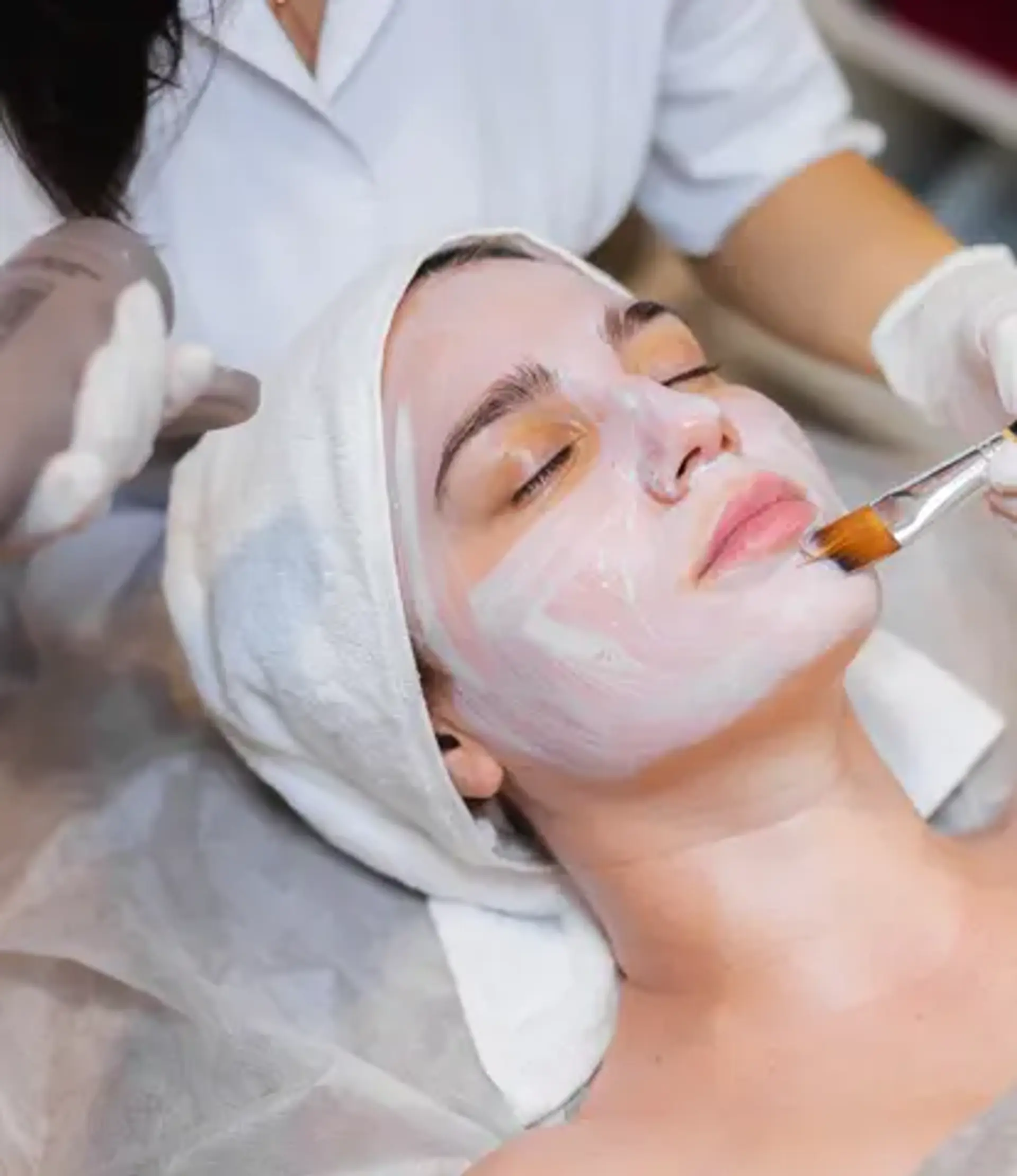 A close-up shot of a woman receiving a facial treatment. A cosmetologist, wearing white gloves, applies a white mask to the woman's face with a brush. The woman has her eyes closed and a white towel wrapped around her head. The setting appears to be a spa or beauty salon.