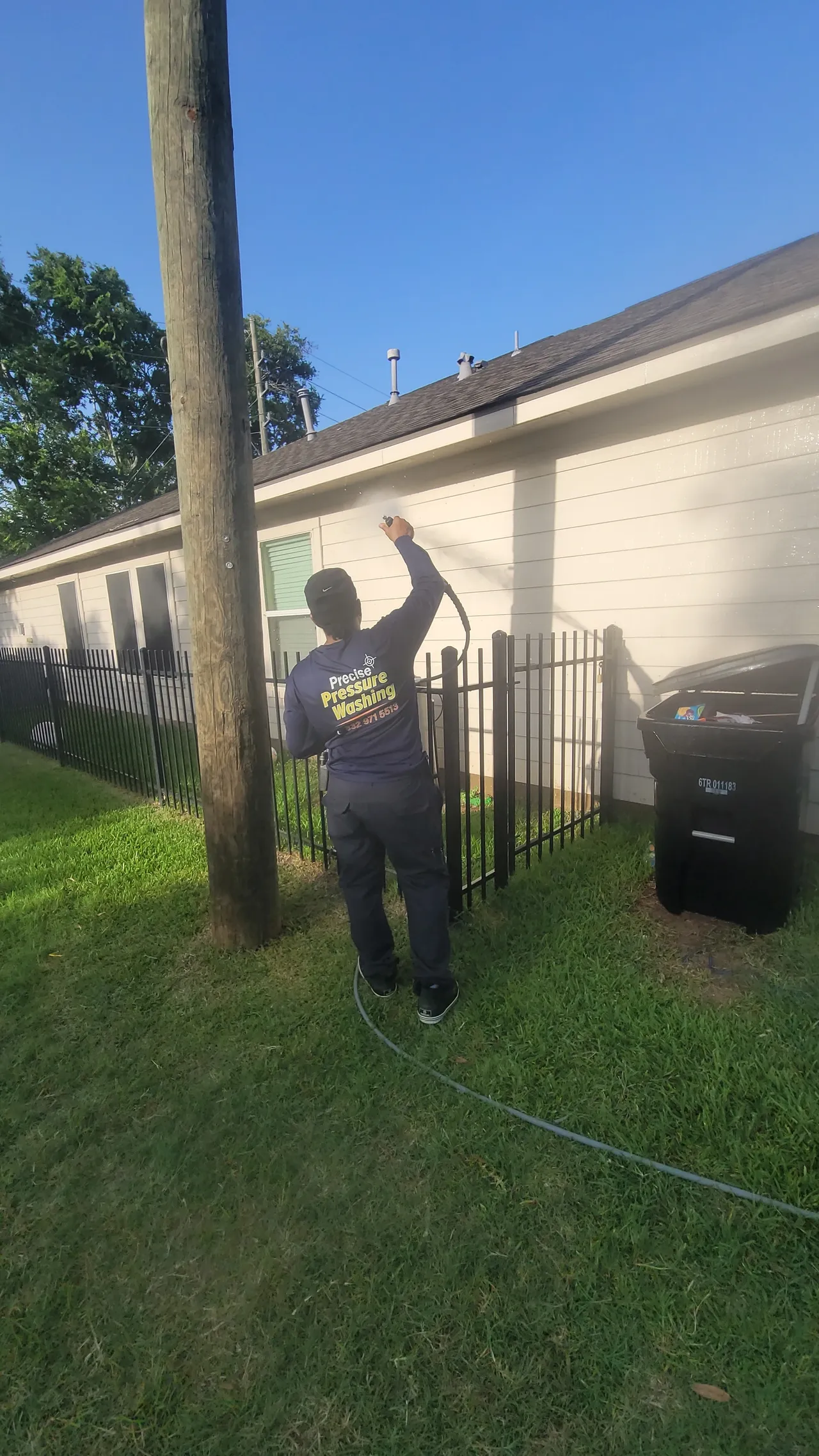 A person pressure washing the exterior of a house on a sunny day. The person is wearing a navy blue long-sleeved shirt with yellow lettering, dark pants, and a black baseball cap. They are holding a pressure washer wand and spraying water on the siding of the house. A black metal fence is visible in the foreground, and a black trash can is to the right. The house has beige siding and several windows. A wooden utility pole stands to the left of the frame. The sky is clear and blue, with green grass and trees in the background.