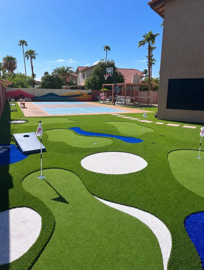 A backyard transformed into an entertainment space featuring a miniature golf course, a basketball court, and a cornhole setup. The artificial turf of the mini-golf course is shaped with white sand traps and blue water features, complete with flagpoles and holes. Adjacent to it is a colorful basketball court with a hoop. The area is surrounded by palm trees and residential buildings, under a clear blue sky.