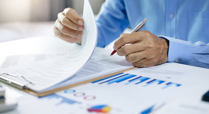 Man writing on one of several documents splayed in front of him on a table.
