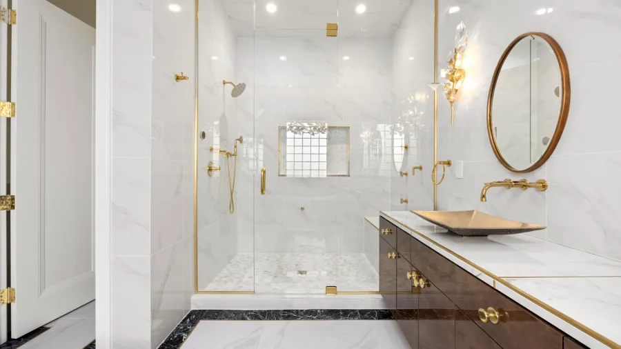 A luxurious bathroom featuring a spacious glass-enclosed shower with marble tile, a modern vanity with a gold geometric sink, a large oval mirror, and gold-toned fixtures. The walls and floor are finished with elegant white marble.
