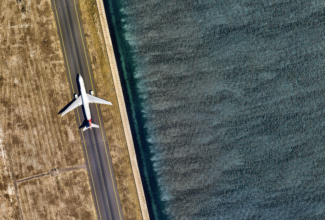 Aerial view of an airplane on a runway beside a calm, textured body of water, bordered by a grassy field, conveying a serene, balanced landscape.