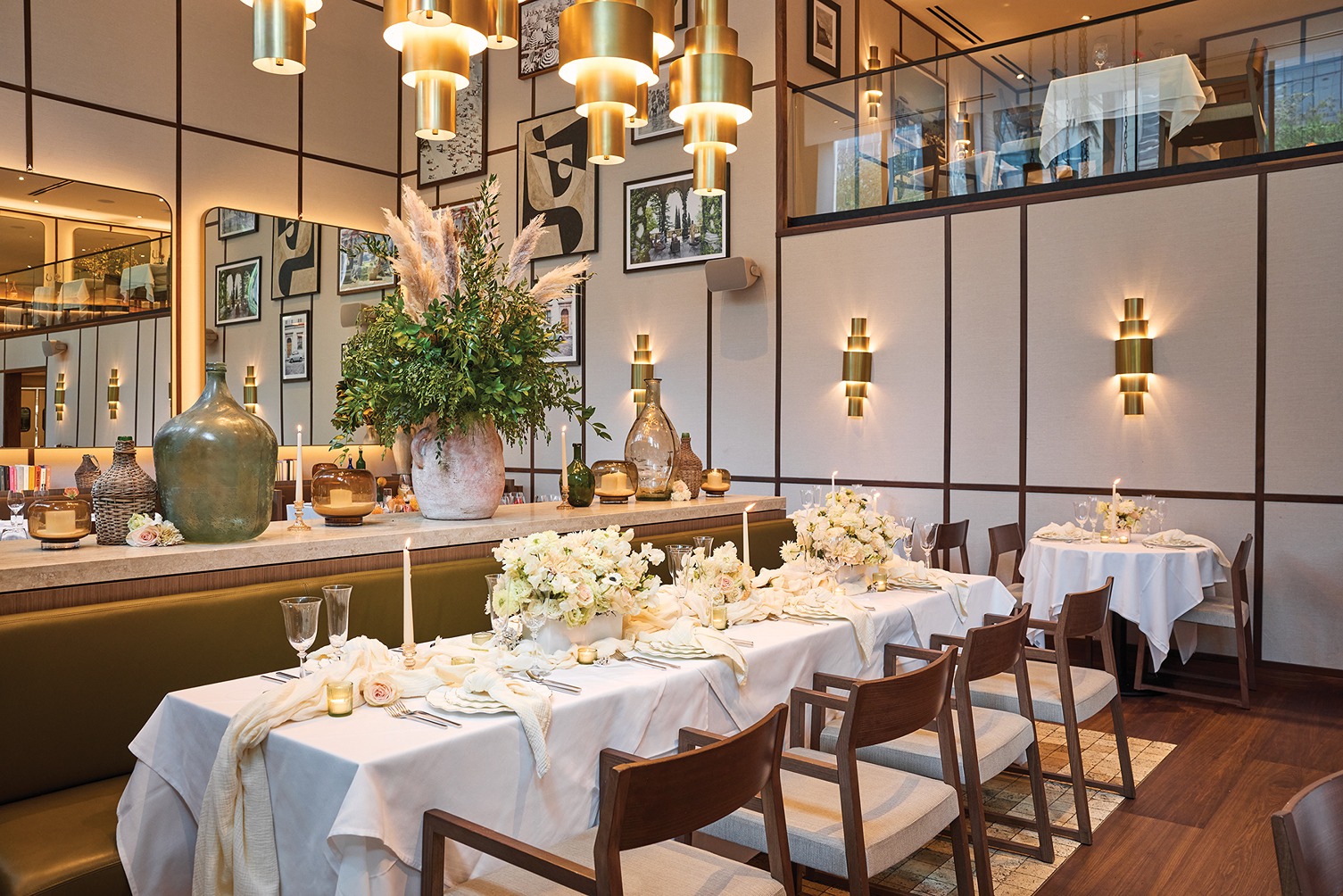A long banquet table prepared for a special event, featuring sophisticated white linens, gold tiered chandeliers, and a gallery wall of art in a modern Italian setting.