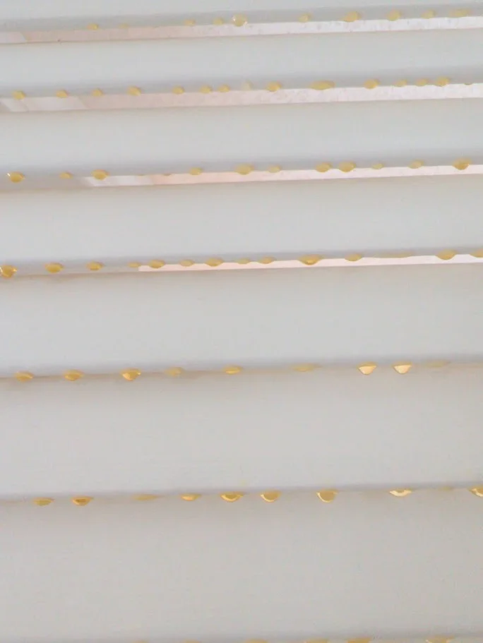 A close-up shot of white horizontal blinds with small, golden-yellow droplets of liquid on each slat.