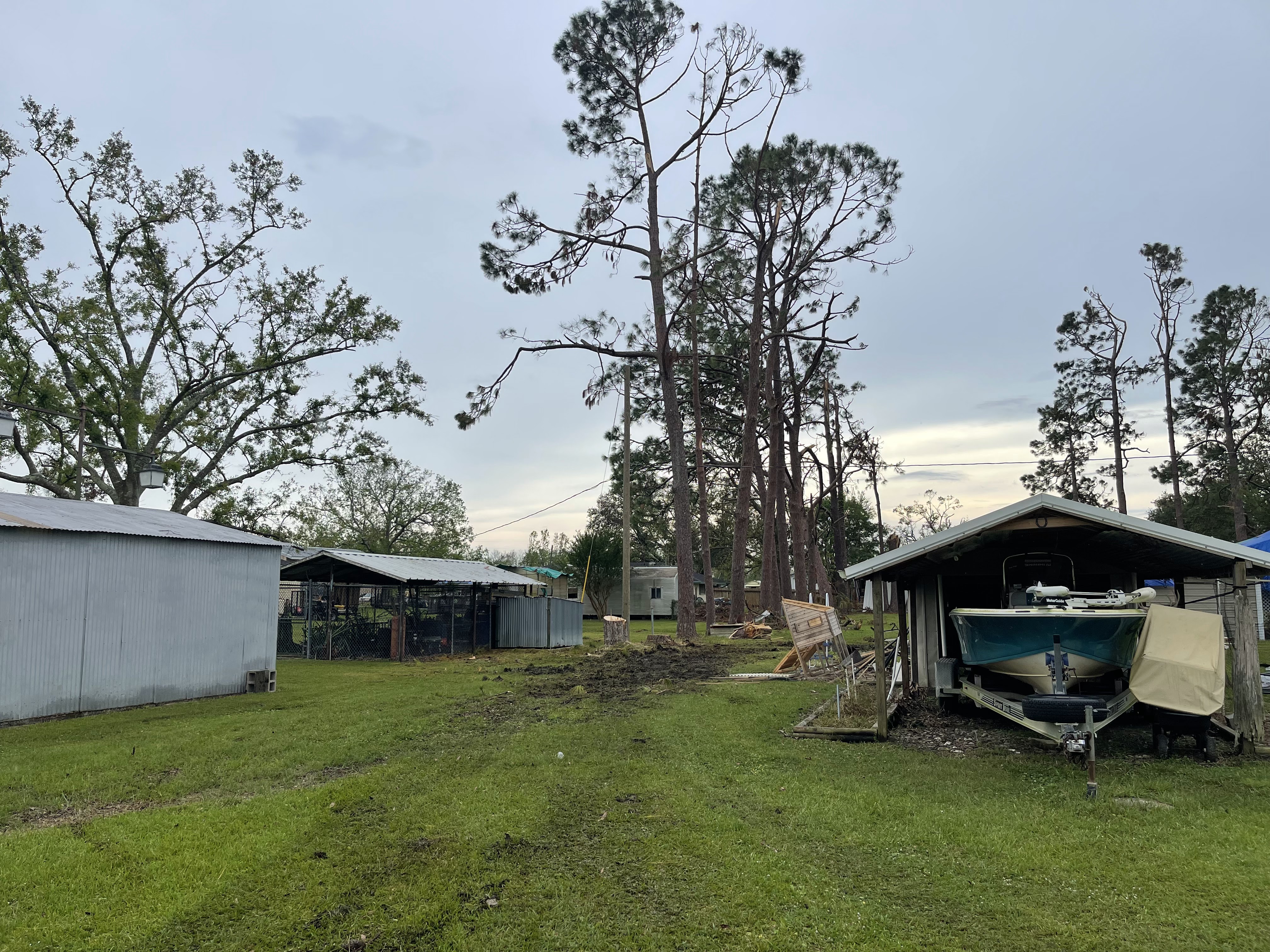 A cloudy sky hangs over a grassy yard with several structures, including a boat on a trailer parked under a covered shelter. Debris is scattered across the yard, suggesting recent damage or cleanup. Tall pine trees dominate the background.