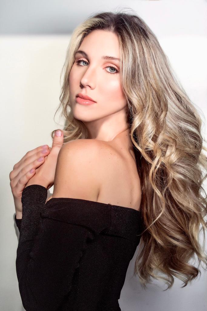 A woman with blonde, wavy hair and blue eyes is shown from the shoulders up, wearing a black off-the-shoulder top. Her left hand is touching her right shoulder. The background is a plain, off-white wall.