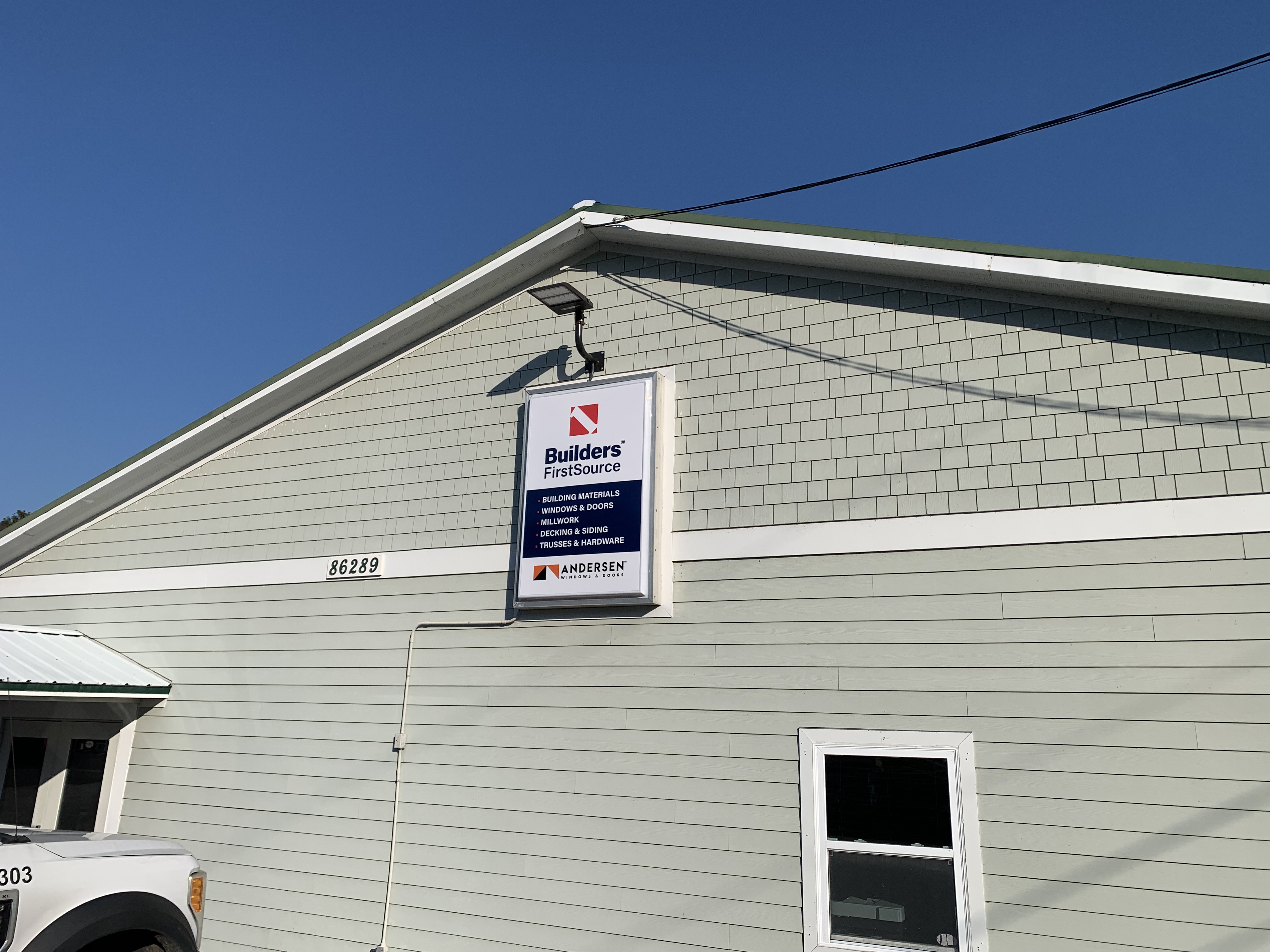 he side view of a beige building with green trim is shown, featuring a sign that reads "Builders FirstSource" with a list of services including building materials, windows and doors, millwork, decking, siding, trusses, and hardware. Below the sign is a logo for Andersen windows. The building number "86289" is clearly visible on the side of the building, and part of a white truck is seen in the bottom left corner. The image was taken on a sunny day with a blue sky.