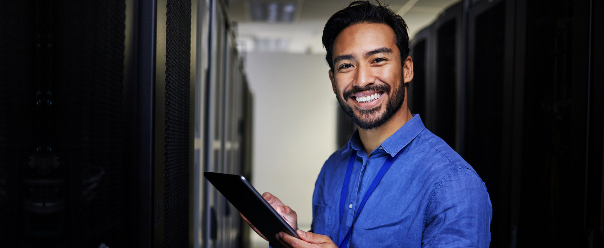 A smiling man with dark hair, a beard, and a blue collared shirt is holding a tablet in a server roo 