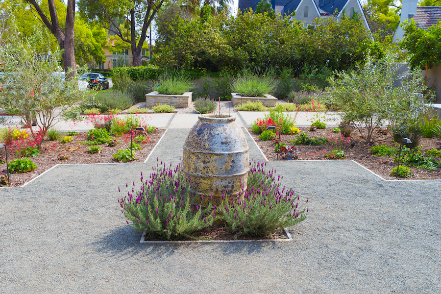 Symmetrical, drought-tolerant garden with a weathered stone fountain, lavender, red flowers, gravel paths, lush greenery, and homes beyond.