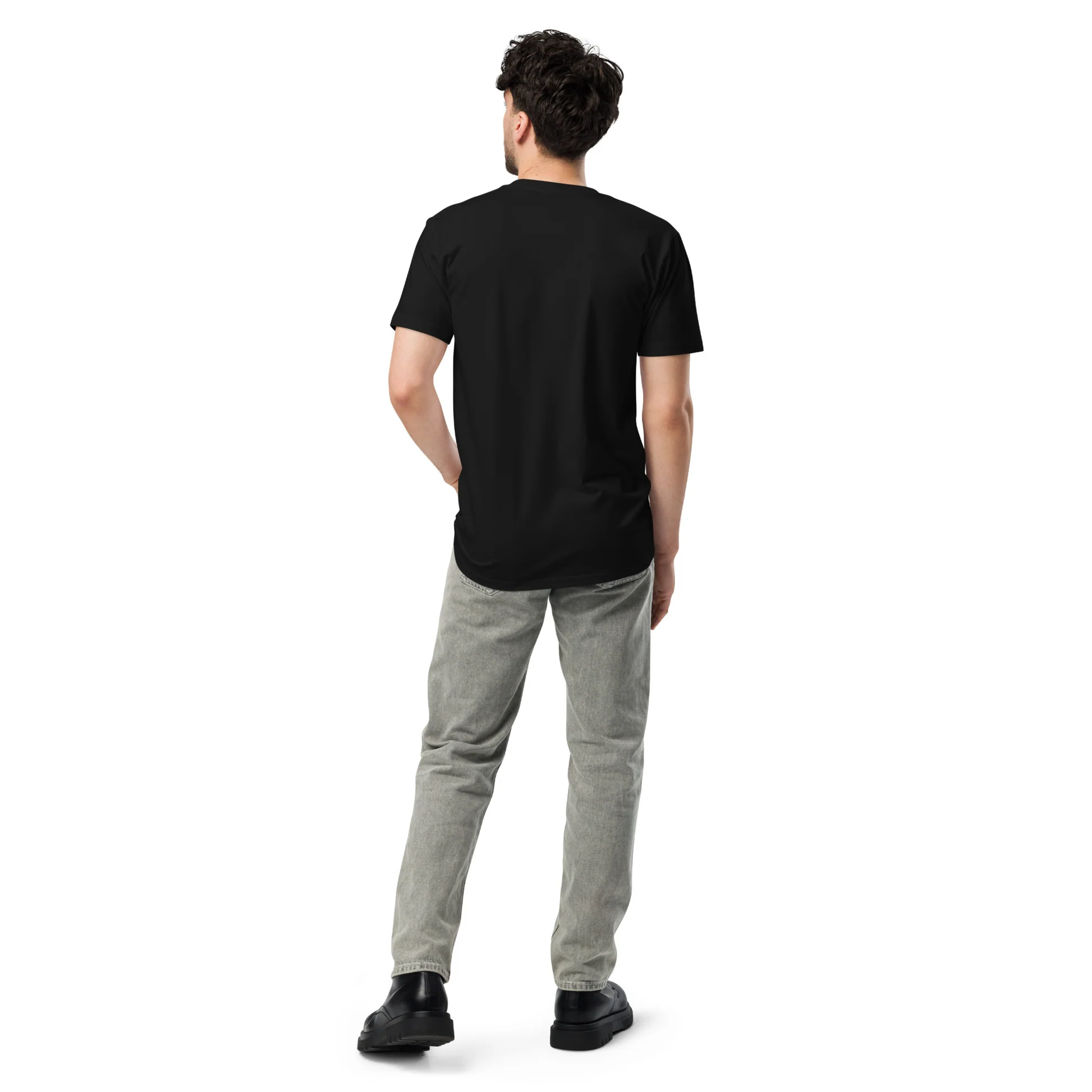 Rear view of a man wearing a plain black t-shirt, grey denim jeans, and black chunky-soled shoes, standing against a white background. His left hand is in his pocket and his head is turned slightly to the left.