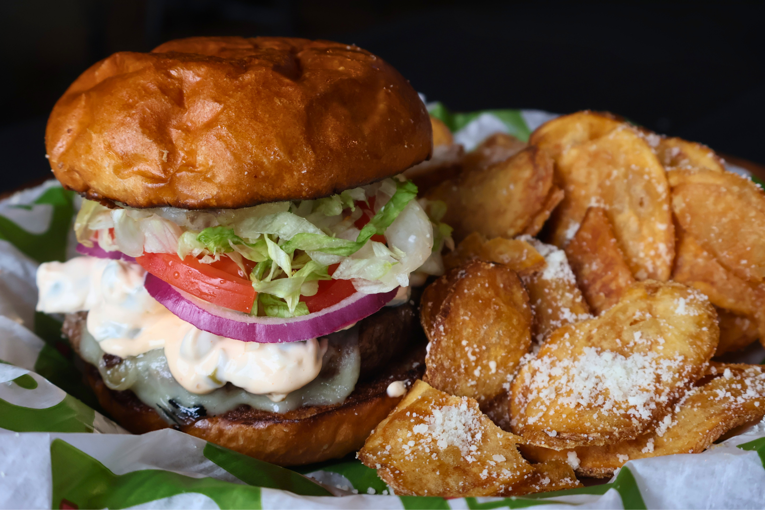 A close-up shot of a juicy burger stacked with fresh vegetables and creamy olive sauce. Beside it, thick-cut, golden-brown potato chips are sprinkled with white cheese, all resting on green patterned paper.