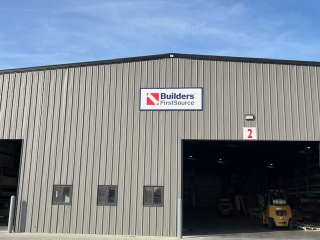 Exterior view of the Builders FirstSource warehouse in Spencer, featuring a large industrial building with a sleek gray metal siding and a gabled roof. The prominent Builders FirstSource logo is displayed at the top center of the building. Below, a wide open bay labeled with the red number '2' provides a glimpse into the warehouse interior, where forklifts and materials are visible. Smaller rectangular windows line the left side, and the bright blue sky with soft, wispy clouds creates a crisp and clear background.