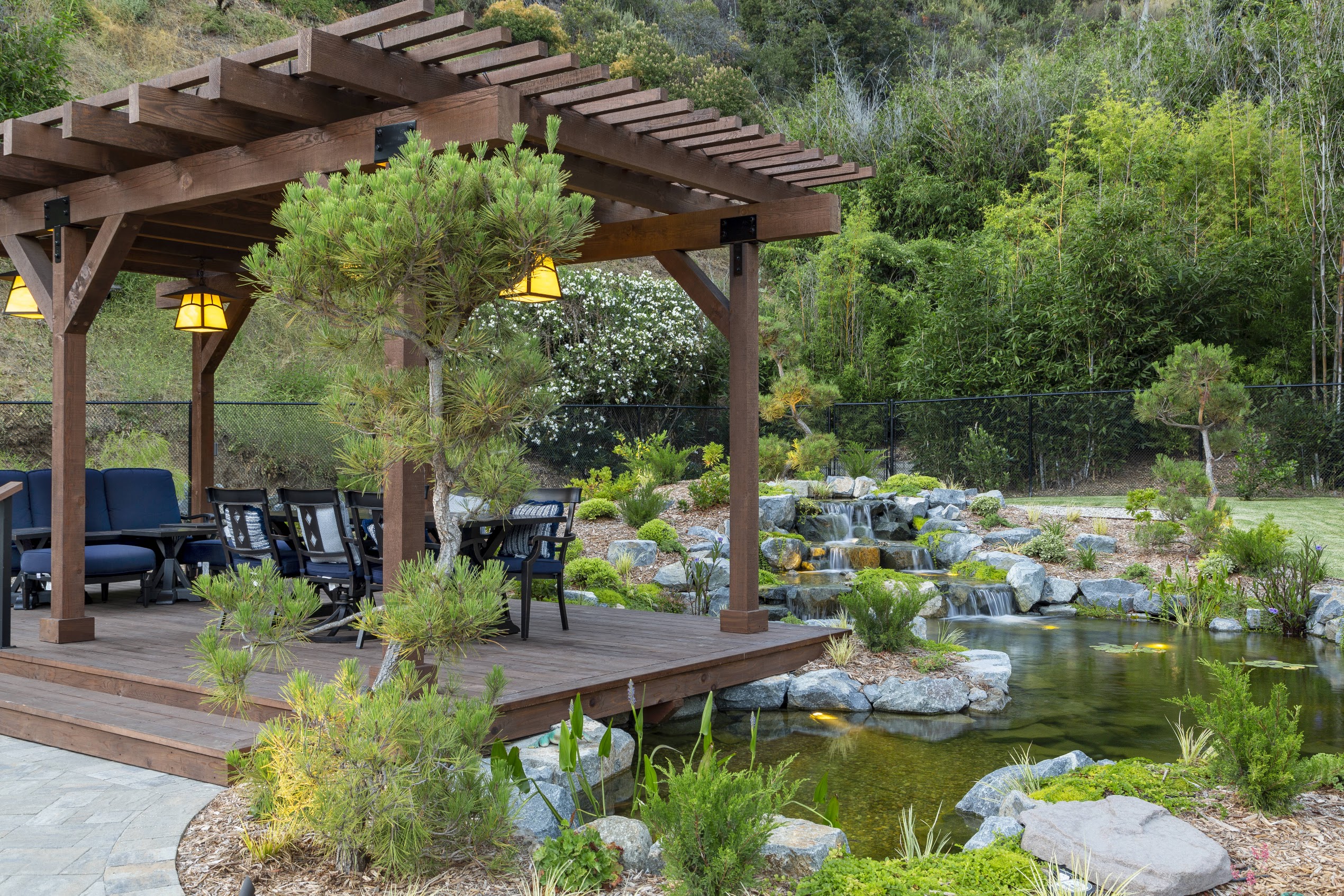 Wooden pergola with seating and glowing lanterns overlooks a landscaped pond, waterfalls, rocks, and lush greenery in a serene garden.