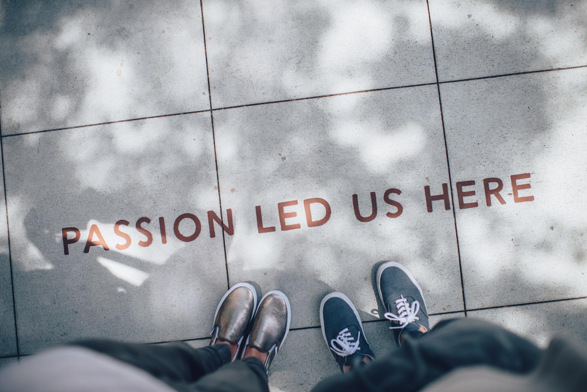 A top-down view of two pairs of feet standing on a sidewalk with the words 'PASSION LED US HERE' etched into the stone. The shadows of trees create a dappled pattern on the ground.