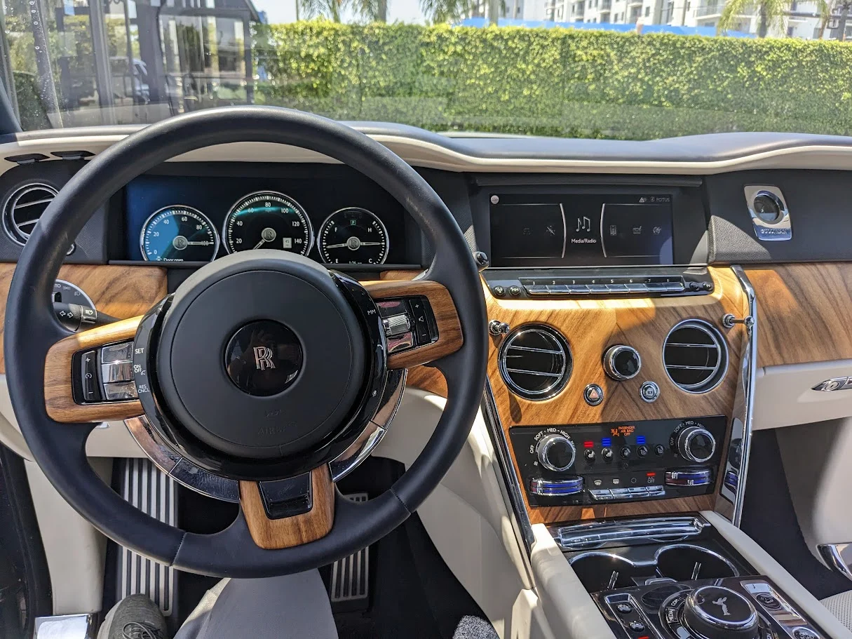 The interior of a luxury car, likely a Rolls-Royce, is shown. The focus is on the dashboard with a leather-wrapped steering wheel featuring the Rolls-Royce emblem, wood trim accents, analog gauges, and a central infotainment screen. The climate control and media system controls are also visible, along with the gear shifter and cup holders.