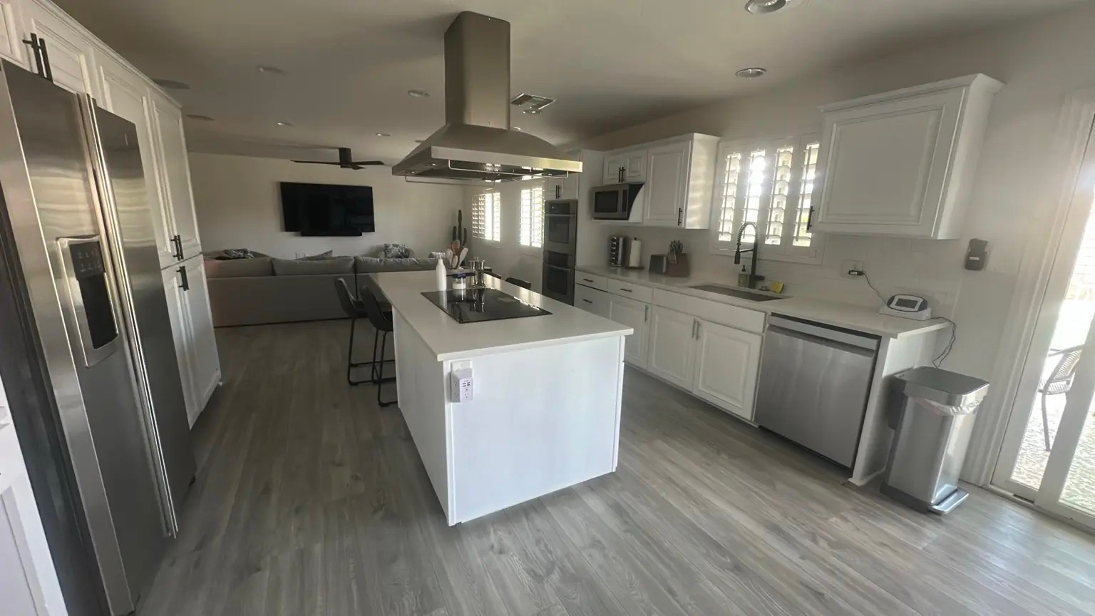 A modern, bright white kitchen flows into an open living space with a large sectional sofa and television. The kitchen features stainless steel appliances, a large island with a cooktop, and ample cabinet space. Sunlight streams in through plantation shutters, illuminating the wood-look tile flooring.