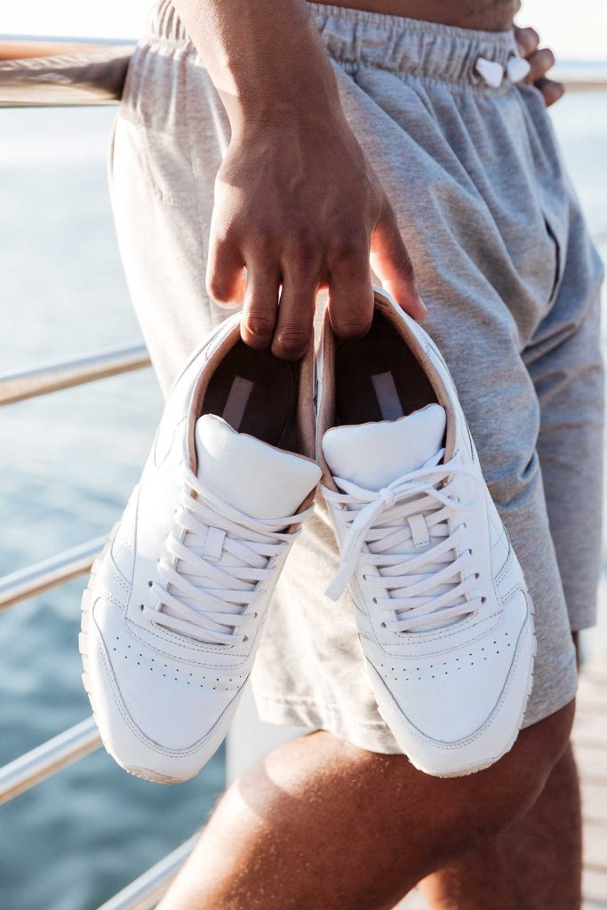 A person holds a pair of white sneakers while wearing gray athletic shorts near a railing with a blurred body of water in the background.