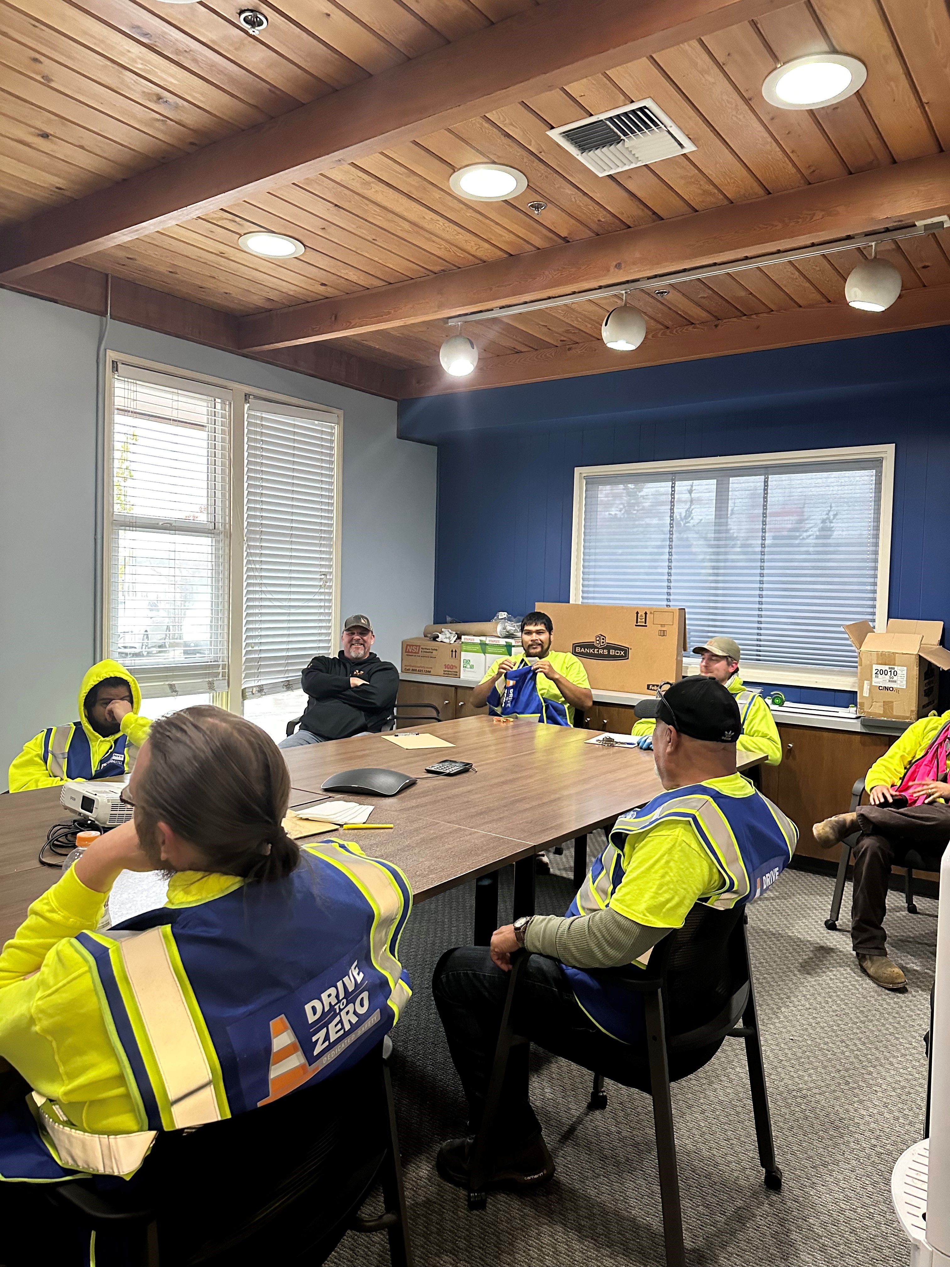 A group of workers is seated around a conference table in a meeting room. Most are wearing high-visibility safety vests with the words "Drive to Zero" printed on the back. The room has wooden ceilings with exposed beams and a large window letting in natural light. The workers appear relaxed, with some in discussion and others listening. Boxes and equipment are stacked along the walls, indicating a workspace or storage area.