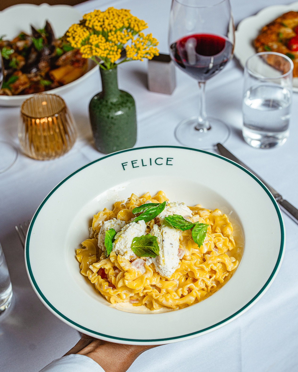 Wavy Mafaldine pasta tossed in a golden sauce and topped with fresh ricotta and basil leaves. The dish is served on a green-rimmed white plate atop a crisp white tablecloth.