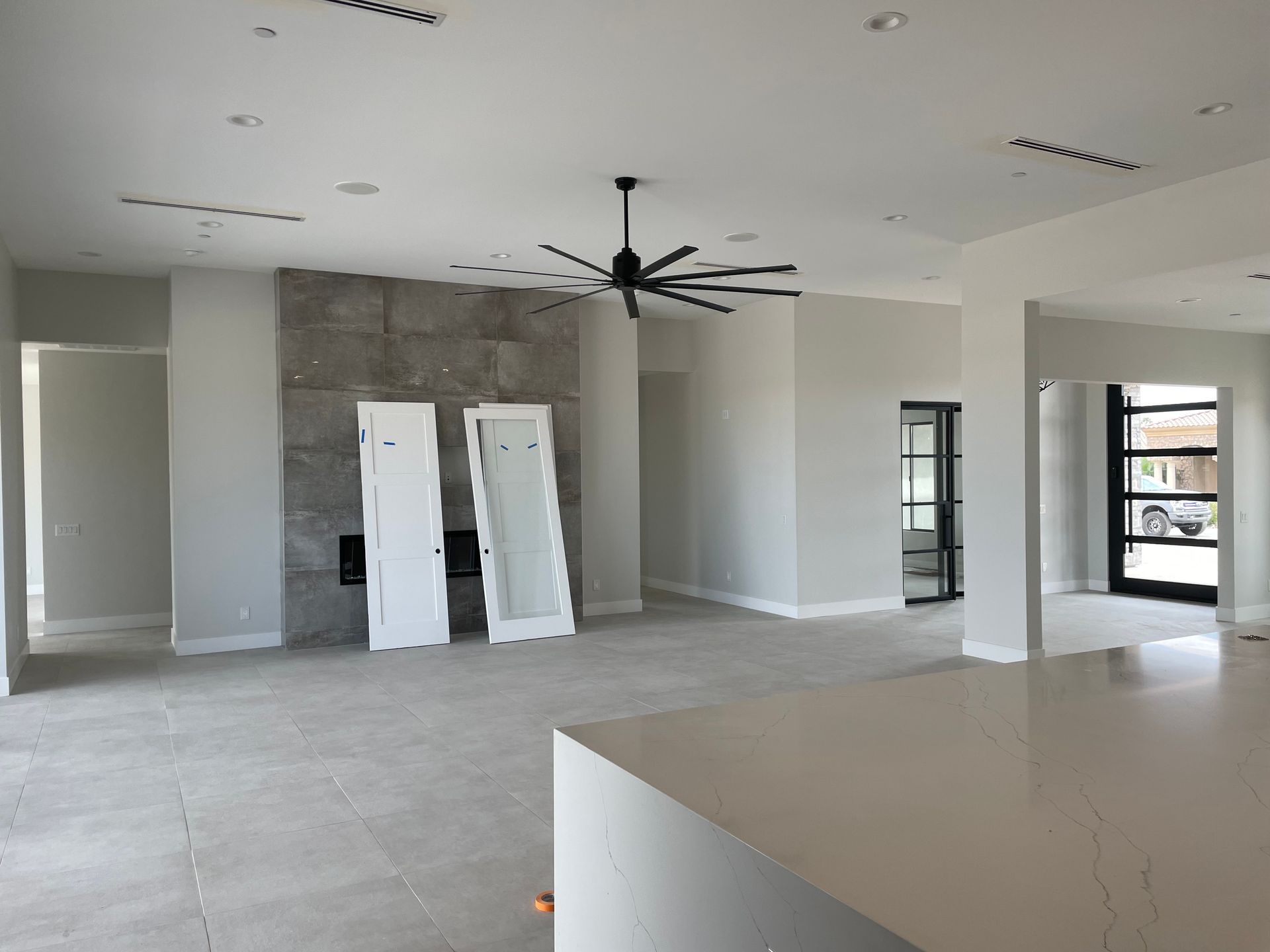 A modern open-concept living space under construction features high ceilings, large ceiling fan, tiled flooring, fireplace wall, and detached doors, showcasing a clean layout with ongoing interior finishing work.
