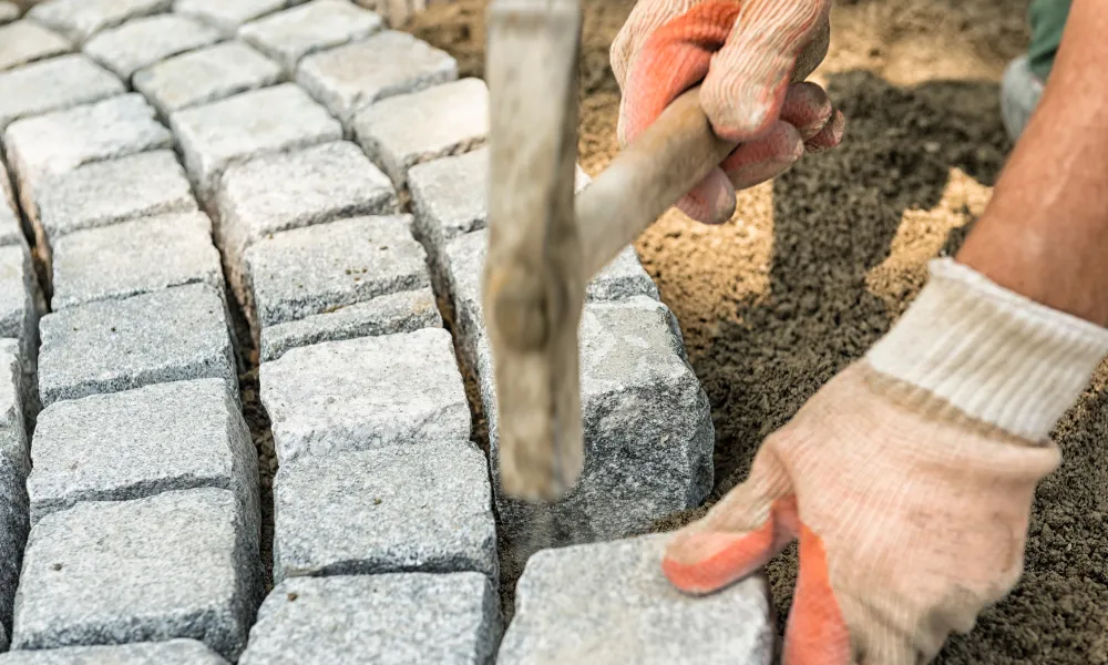 A person wearing orange and white gloves is laying granite cobblestones with a hammer. The cobblestones are arranged in neat rows and are partially embedded in sand or dirt. The focus is on the hands, the hammer, and the stone work in progress.