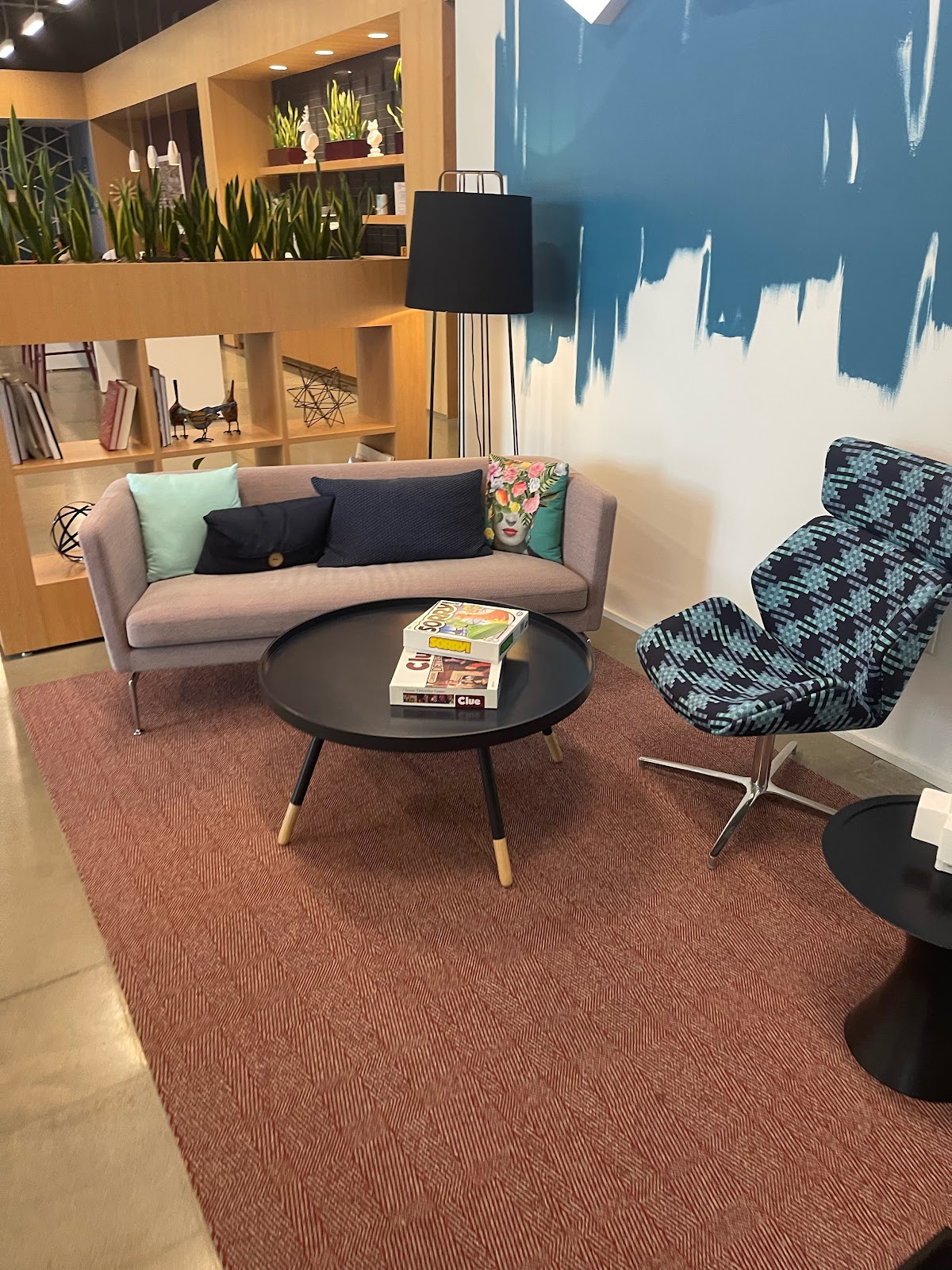 A modern waiting area with a pink sofa, a patterned armchair, and a round coffee table displaying two board games, all set on a red patterned rug.