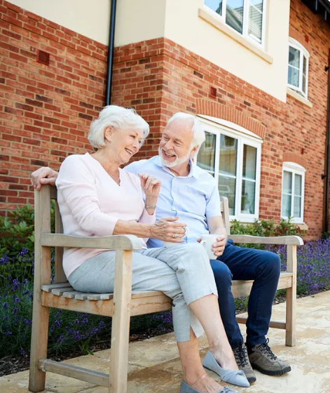 Senior man and woman sitting together on a wooden garden bench outside a modern brick residential building, smiling and holding coffee cups.