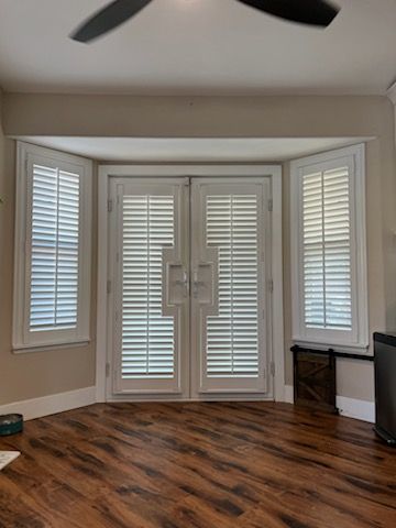 Elegant white plantation shutters expertly fitted to French doors and side windows. The specialized handle cutouts ensure seamless operation while providing a clean, cohesive look that enhances the room's natural light.