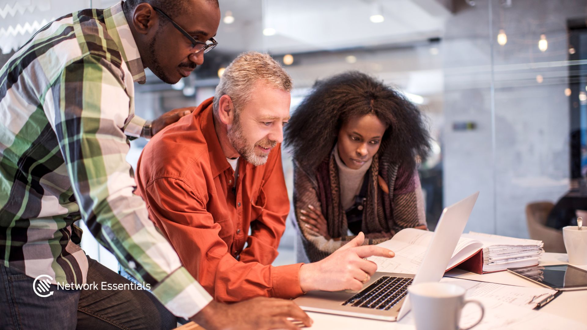 Three professionals, two men and one woman, are gathered around a laptop at a desk, appearing to be in a collaborative work environment. One man, wearing an orange shirt, is pointing at the laptop screen while the other man, in a plaid shirt, leans in to observe. The woman, in a patterned scarf, looks intently at the documents spread across the desk. A logo for "Network Essentials" is visible in the bottom left corner. The scene suggests a business meeting or a work session focused on reviewing information.
