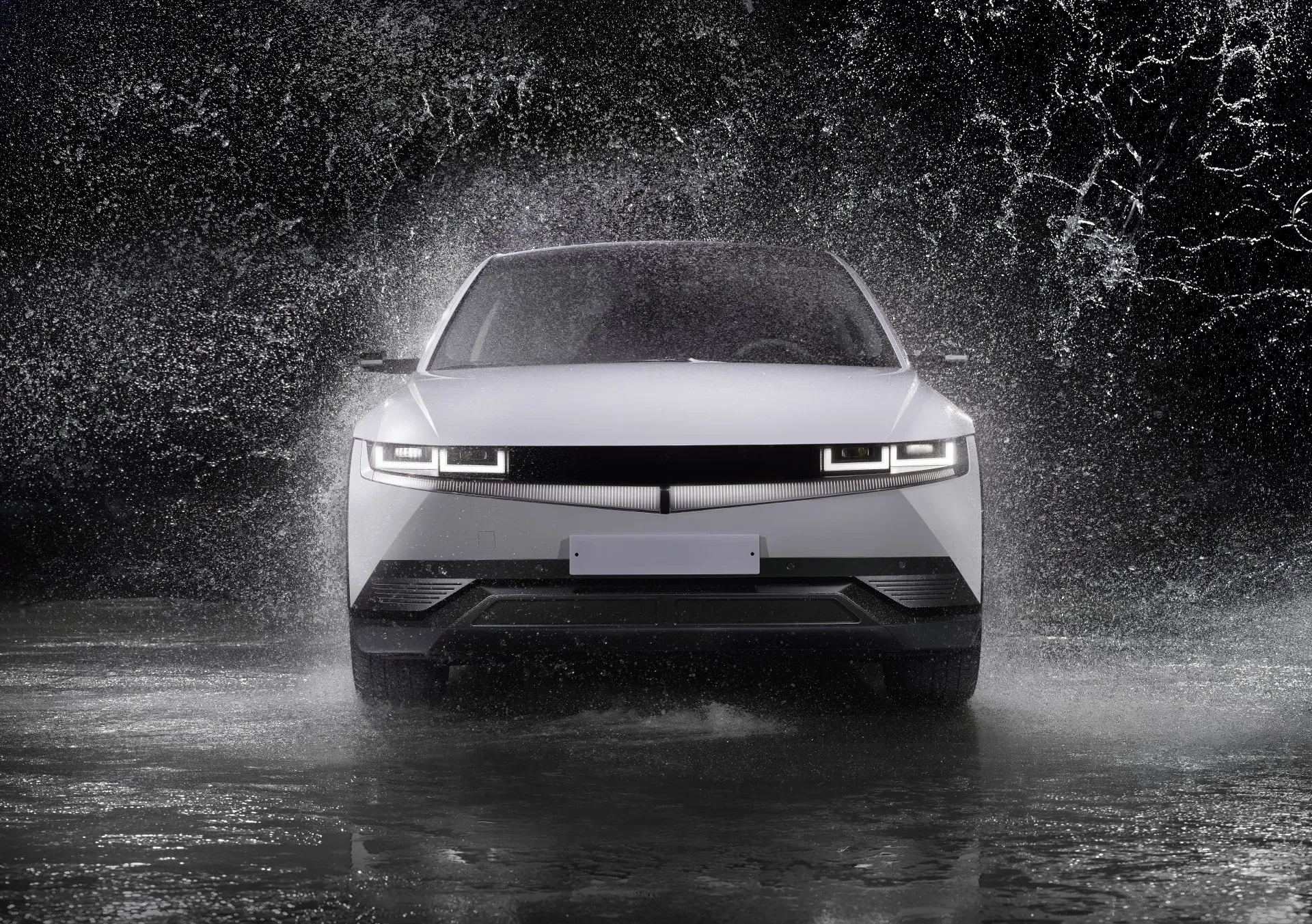 A white Hyundai Ioniq 5 electric car is shown from the front, driving through a splash of water against a black background. The car's distinctive pixelated headlights and taillights are illuminated, and water droplets are frozen in motion, creating a dramatic effect. The wet ground reflects the car and the splashing water, adding to the dynamic scene.