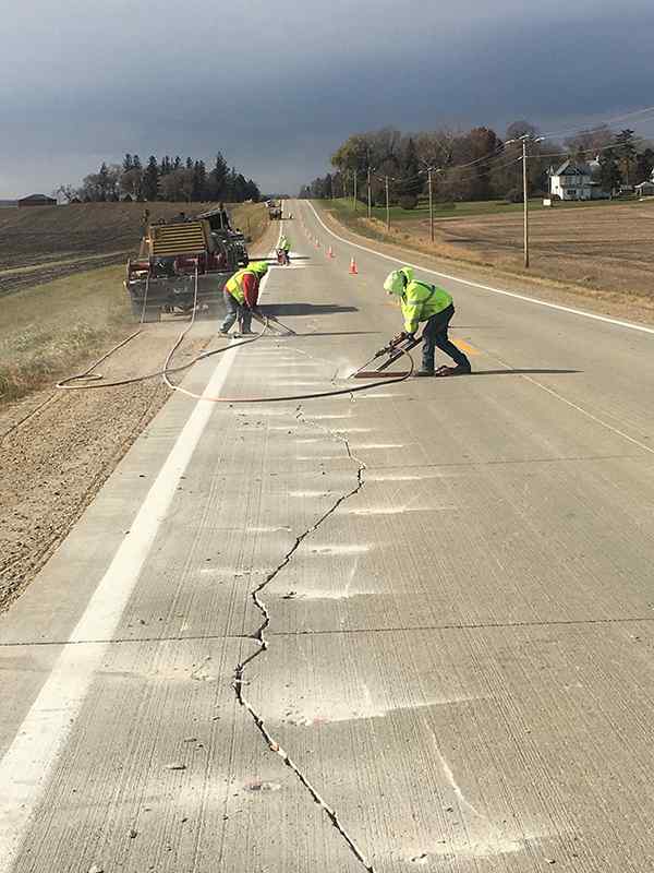Rural highway scene under a dramatic, cloudy sky showing road repair workers actively using handheld pneumatic drills/jackhammers on concrete pavement. In the foreground, a long, jagged crack runs along the lane, with white guidelines marked for repair cuts.