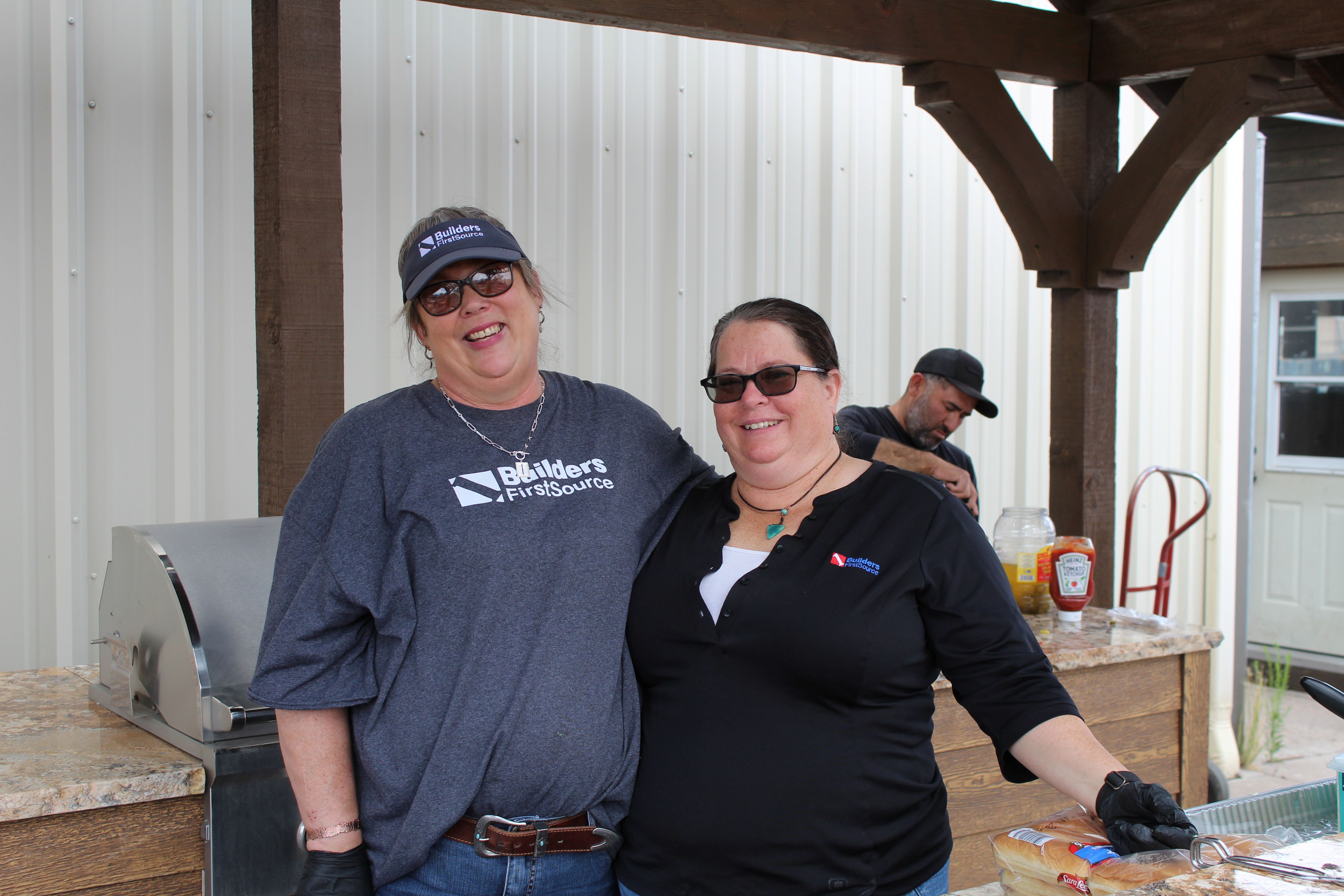 Two Builders FirstSource employees stand together at an outdoor kitchen display during a customer appreciation event, with a grill, food service area, and event activities in the background.