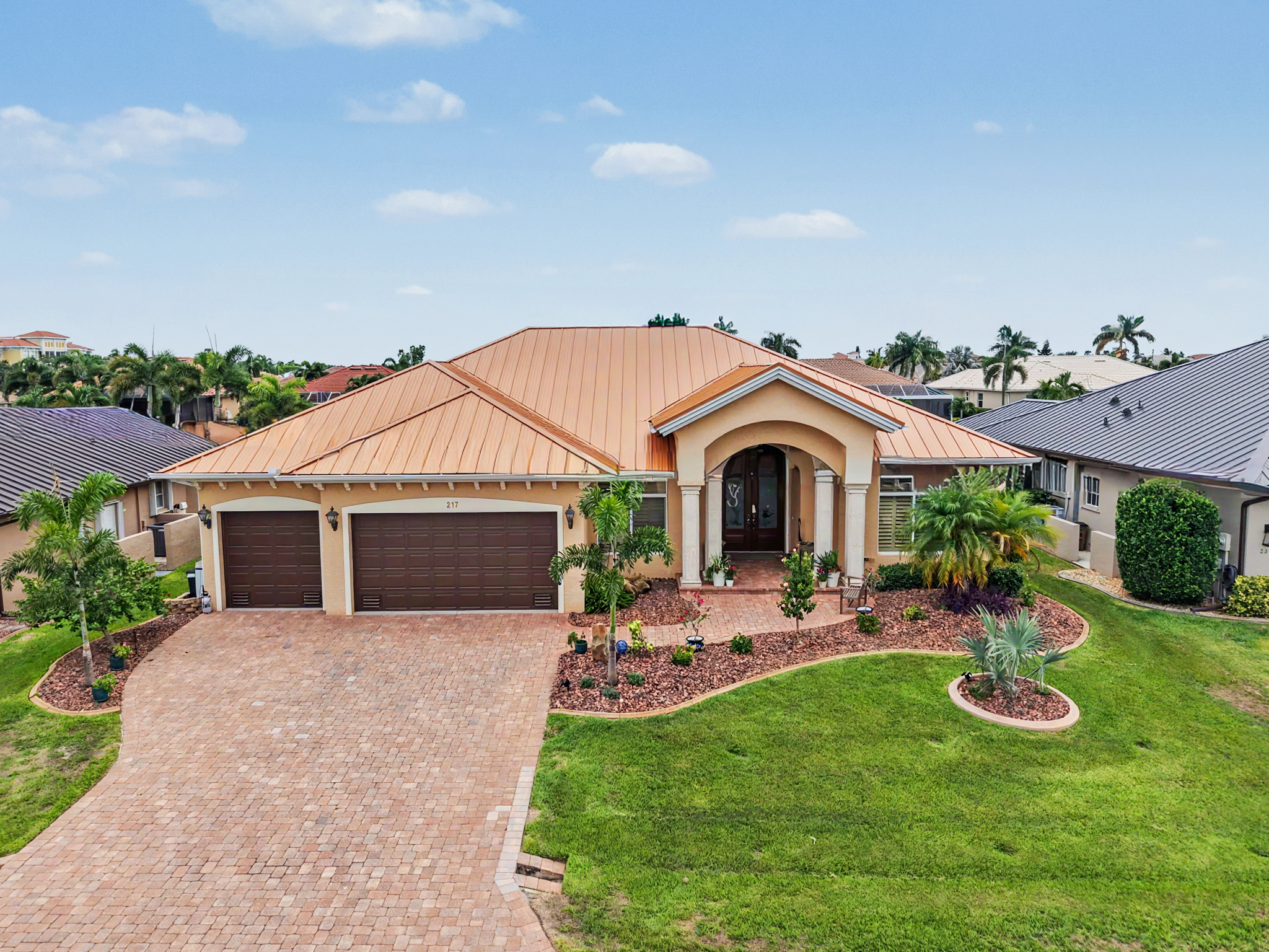 Front drone view of a single-story home with a copper standing seam metal roof, brick driveway, trop 