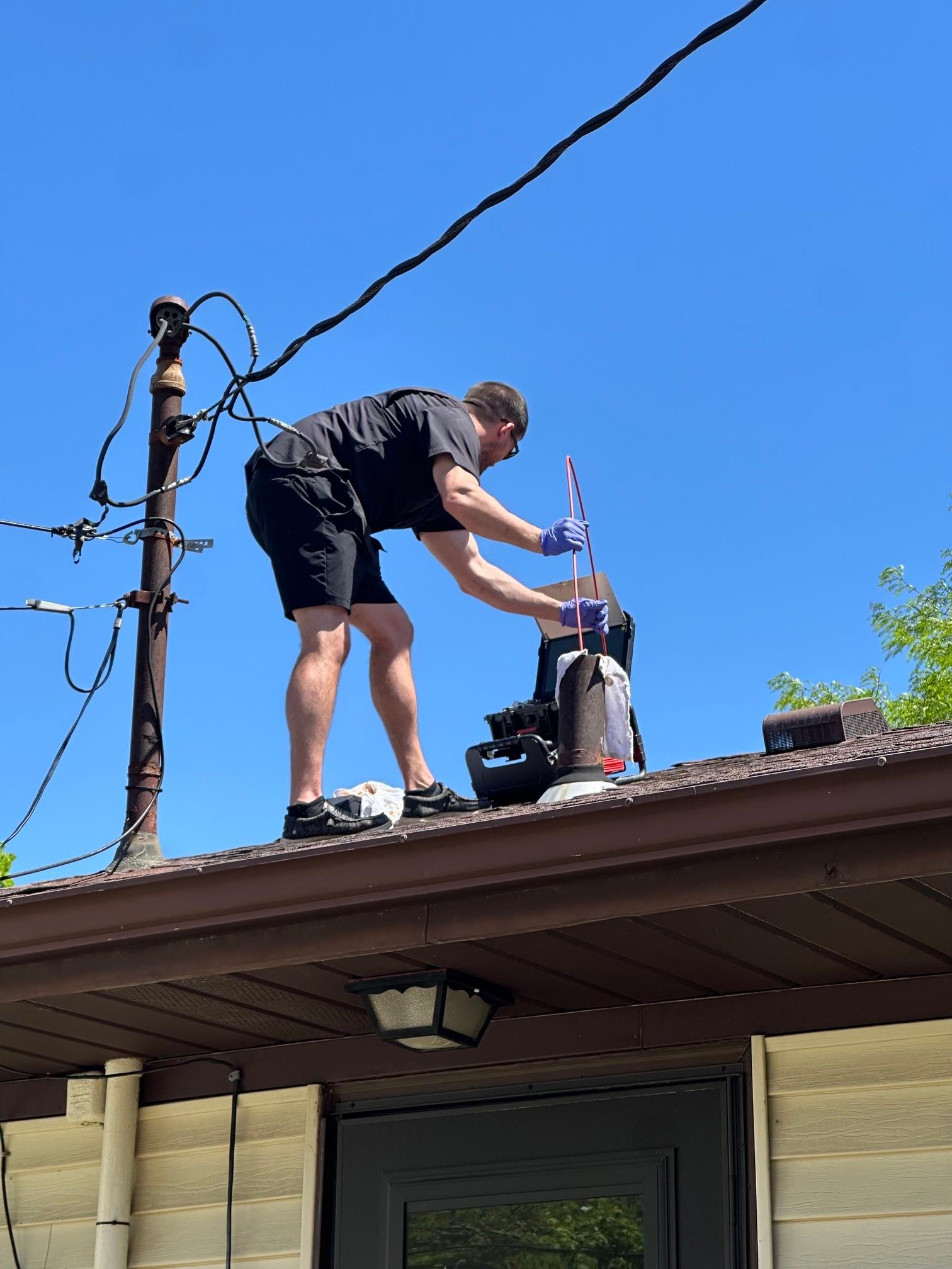 Home inspector feeding a camera line down a roof plumbing vent pipe against a clear blue sky.
