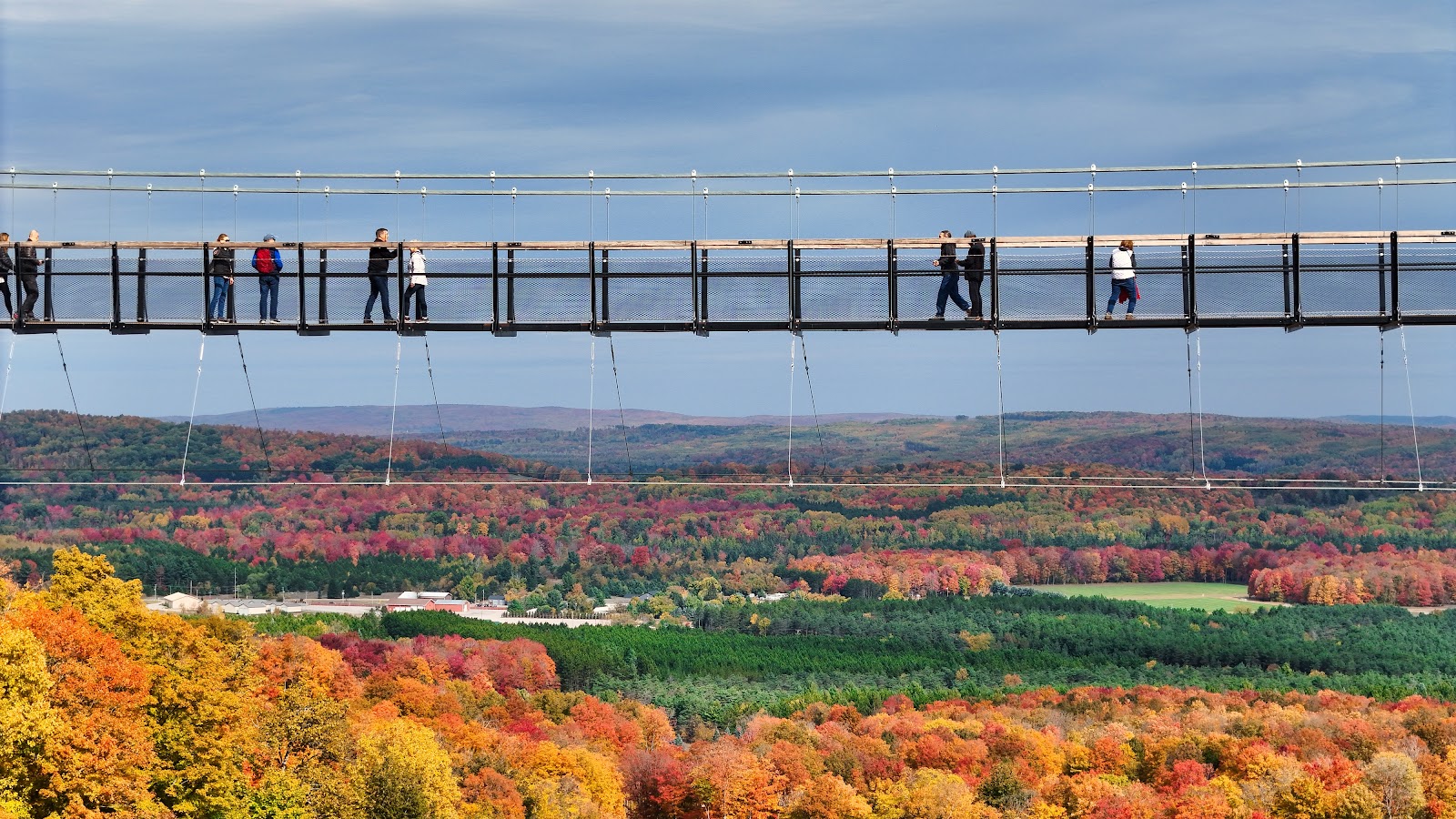 A panoramic view captures a suspension bridge with several people walking across it, set against a backdrop of a dense forest ablaze with autumn colors. The trees display a vibrant tapestry of red, orange, and yellow leaves, with hints of green interspersed throughout. The sky above is a soft blue with scattered clouds.