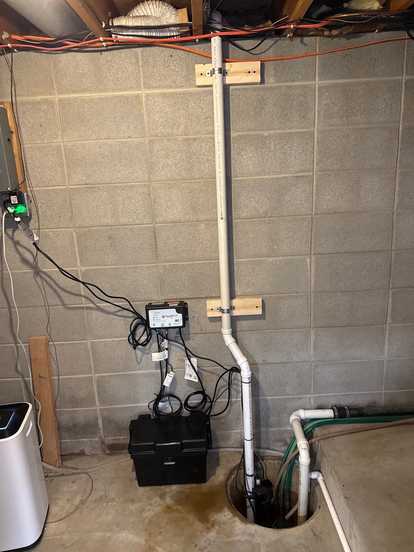Basement sump pump system installed beside a concrete block wall, featuring vertical PVC discharge piping, electrical wiring, control equipment, hoses, exposed ceiling framing, and an open sump pit below nearby.