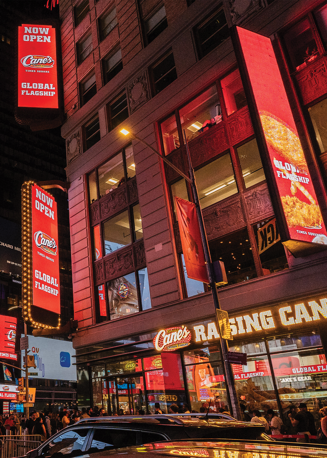 A low-angle nighttime view of the Raising Cane's Global Flagship store in Times Square. The multi-story building is illuminated by massive, bright red digital billboards reading "Now Open" and displaying images of chicken fingers. A busy crowd of pedestrians lines the sidewalk beneath the glowing "Raising Cane's" storefront sign, with the roofs of cars visible in the immediate foreground.