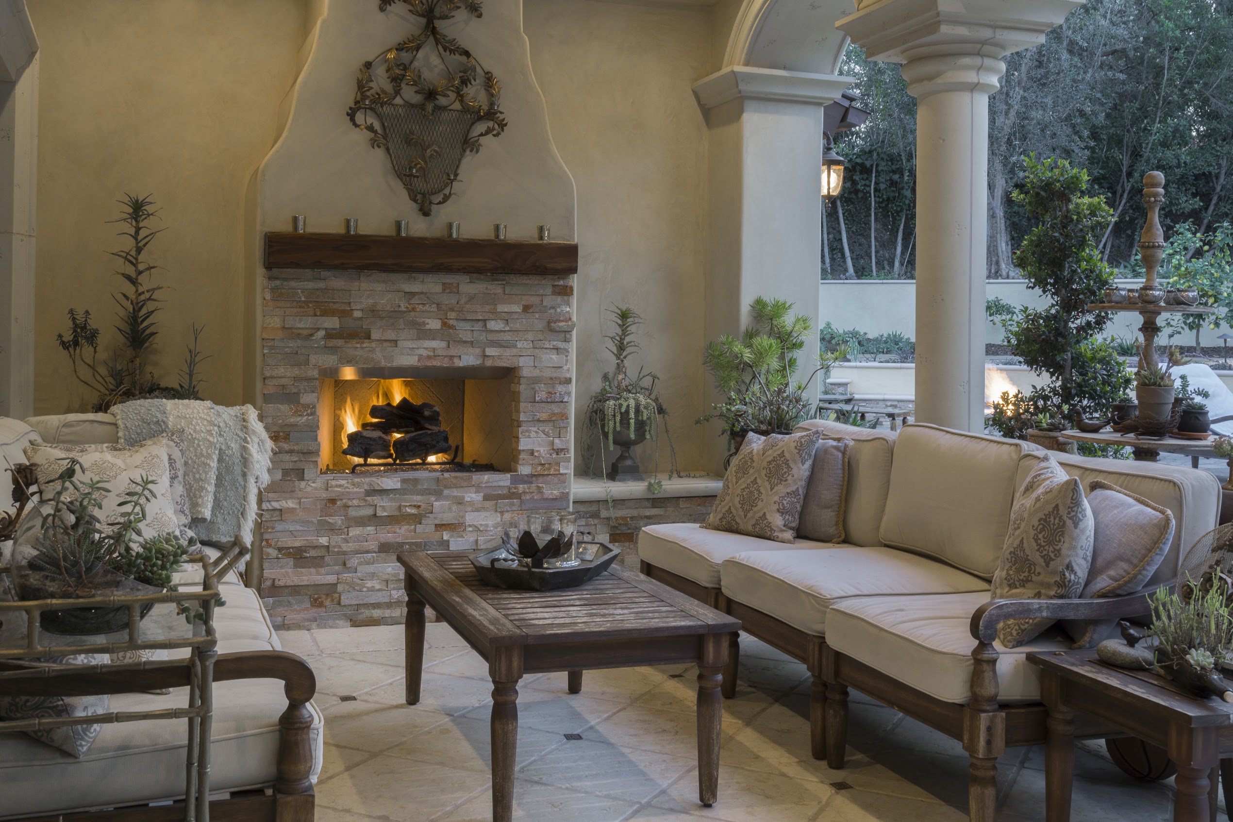 Elegant Mediterranean living room with a glowing stone fireplace, cream sofas, wooden coffee table, arched columns, and lush plants.