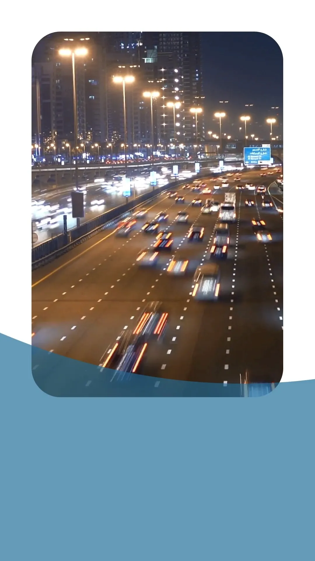 A night-time view of a busy highway with streaks of light from moving cars and illuminated by streetlights and the lights of tall buildings in the background. The image captures the dynamic movement of traffic in an urban environment.