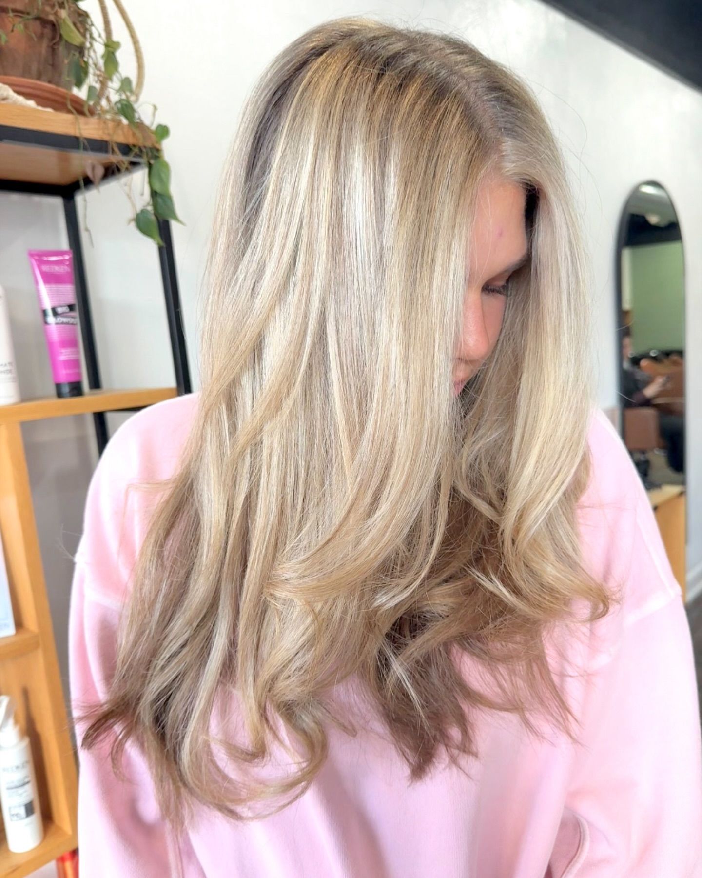 Woman with long layered blonde hair styled in soft waves, photographed indoors in a salon with shelves and styling products.