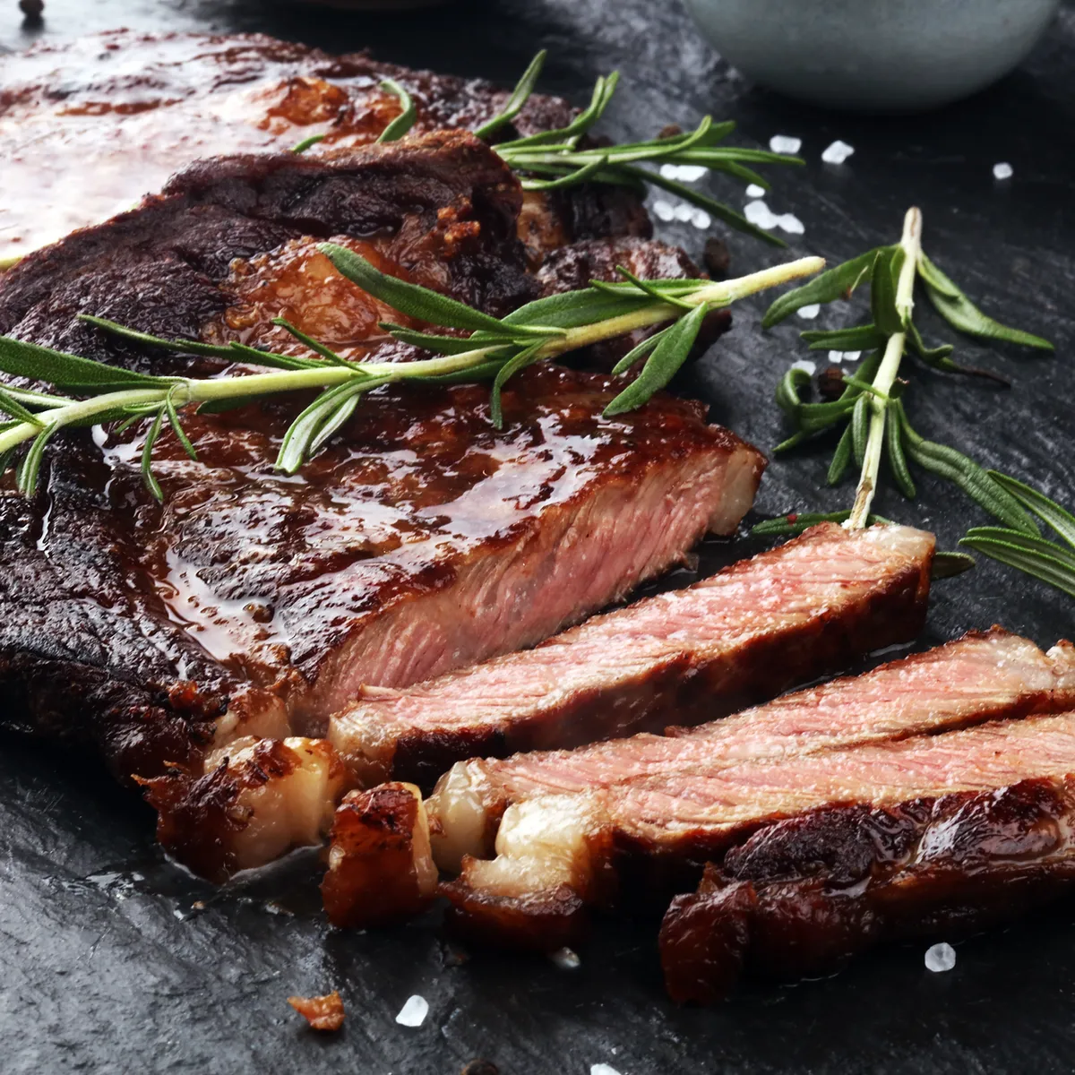 A close-up, artfully arranged shot showcases a thick, juicy, and perfectly cooked ribeye steak. The steak is sliced, revealing a beautiful pink interior with a well-seared, dark brown crust. Fresh sprigs of rosemary are artfully placed over the steak, adding a touch of green and aromatic appeal. Coarse sea salt and black peppercorns are scattered across the dark, textured surface, likely a slate or stone platter, enhancing the rustic and gourmet presentation. The lighting is dramatic, highlighting the rich textures and glossy surface of the meat. In the background, a blurred grey bowl suggests a dining setting.