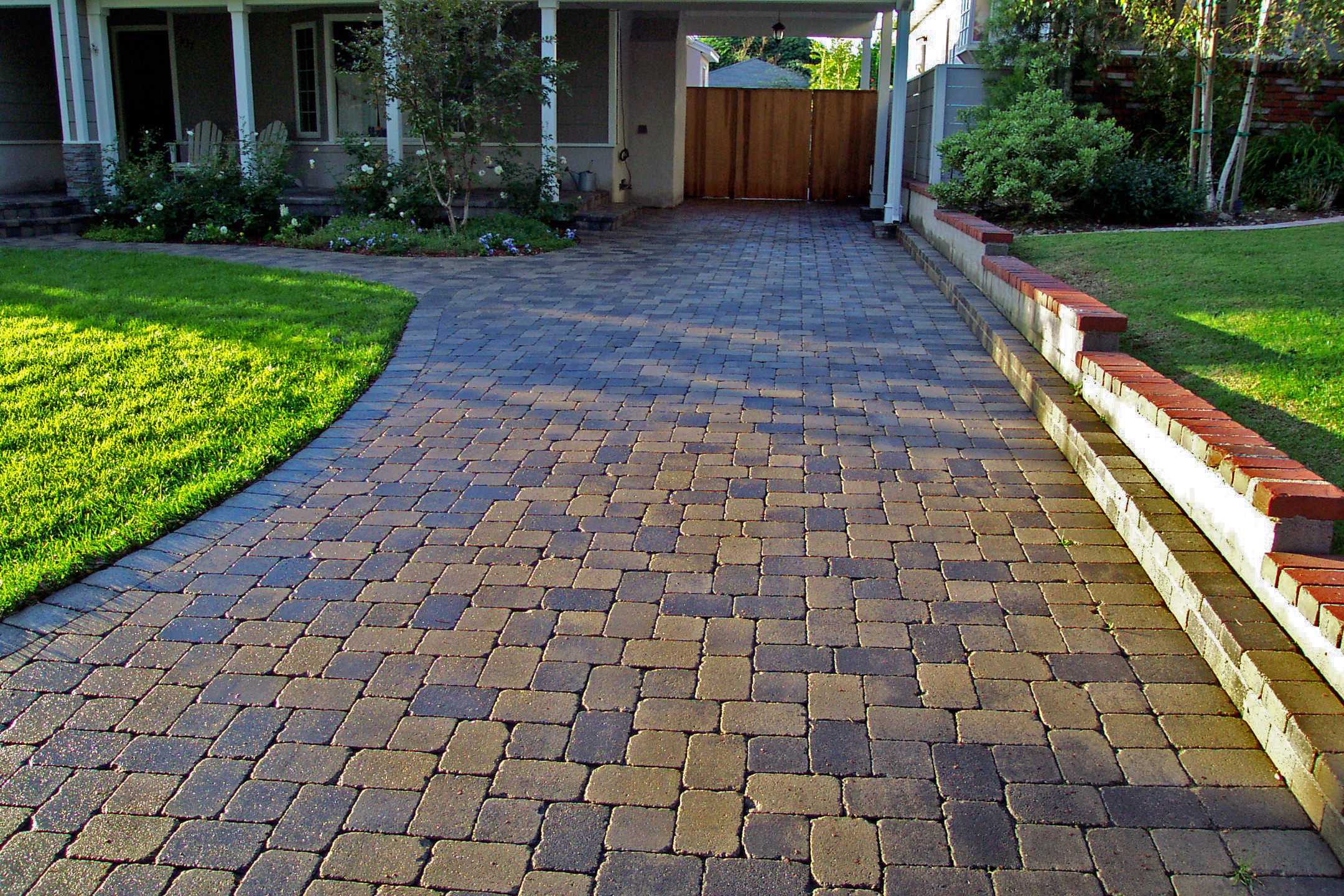 Brick-paver driveway leads to a covered home entrance and wooden gate, bordered by bright green lawns and a low red-brick wall.