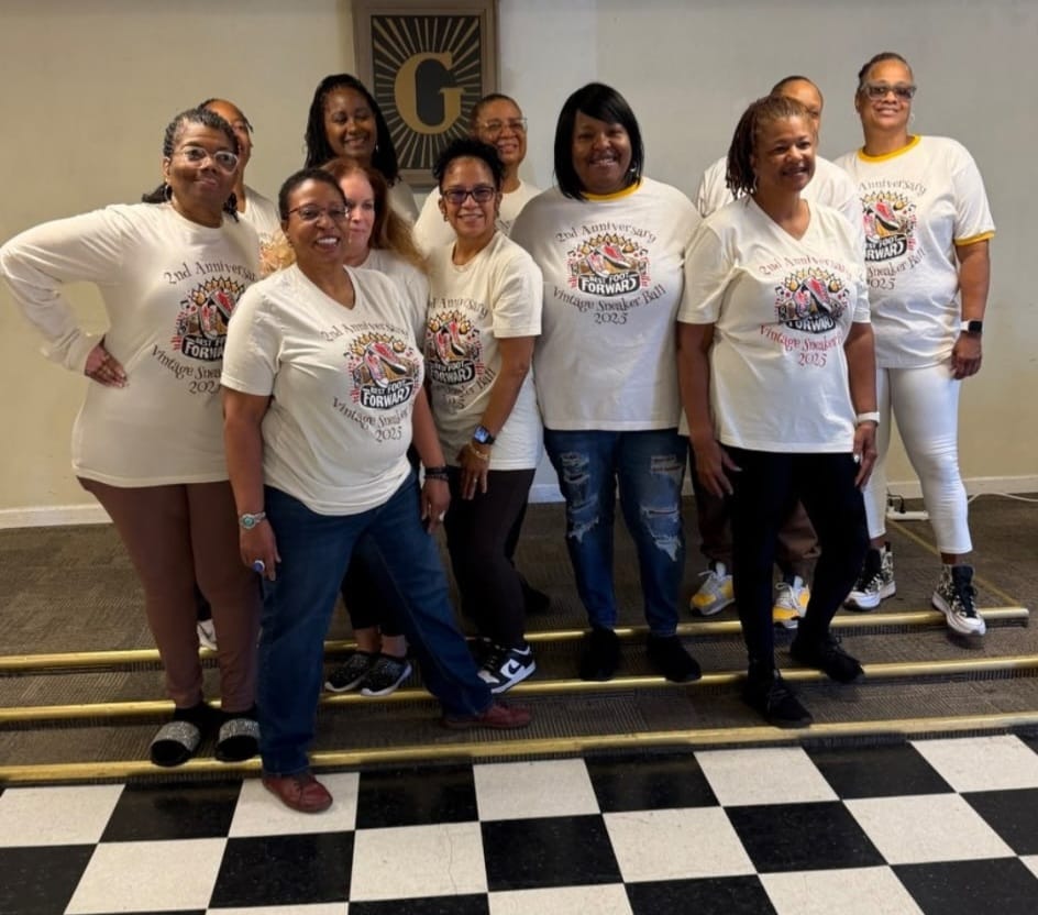 A group of ten women, all wearing matching long-sleeved t-shirts, pose for a photo. The t-shirts are off-white with a graphic design featuring the words "2nd Anniversary", "BEST FOOT FORWARD", and "Vintage Sneaker Ball 2023". The women are standing on a set of stairs with a black and white checkered pattern on the steps. Behind them, a framed picture with a large "G" is mounted on the wall. The overall scene appears to be an indoor event or gathering.