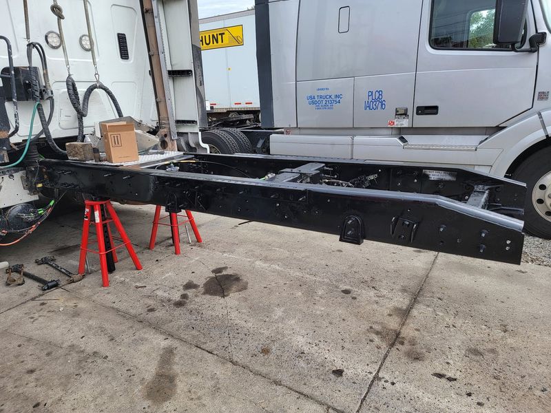 A mechanic stands next to a long, black-painted heavy-duty truck chassis frame that has been strippe 