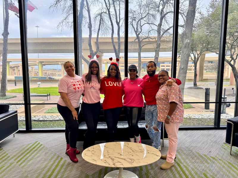 Six people, three men and three women, are posing for a photo in an indoor setting with large windows overlooking an urban landscape. They are all dressed in shades of pink and red, with some wearing Valentine's Day-themed accessories like heart headbands. The group appears to be colleagues or friends celebrating an occasion.